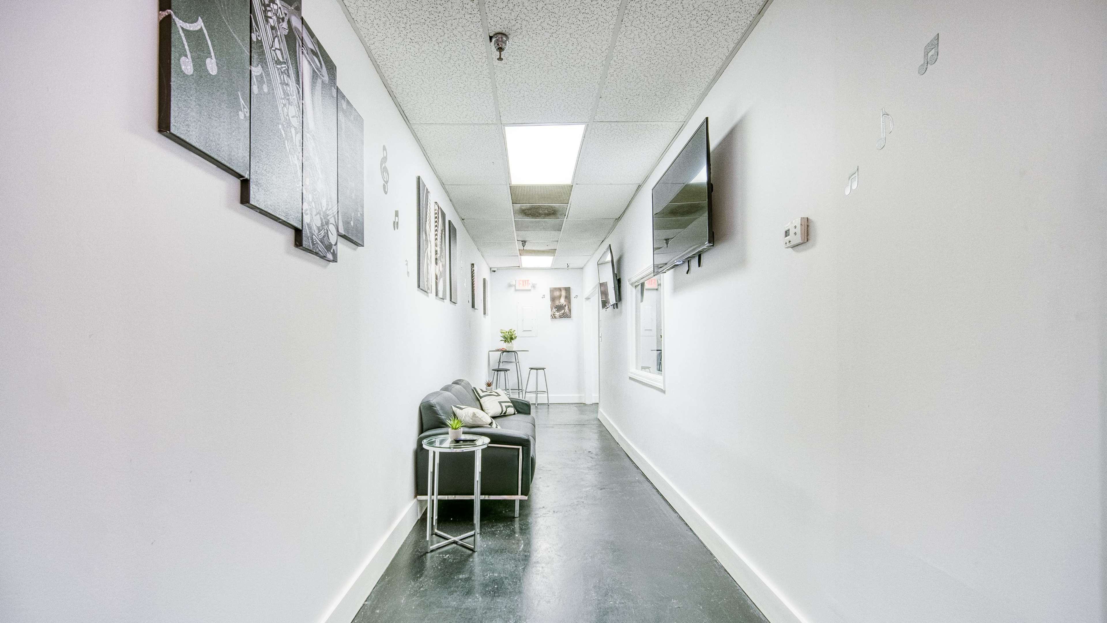 A long, narrow hallway with a black couch and a small table, framed artwork on the walls, and a television mounted on one side.