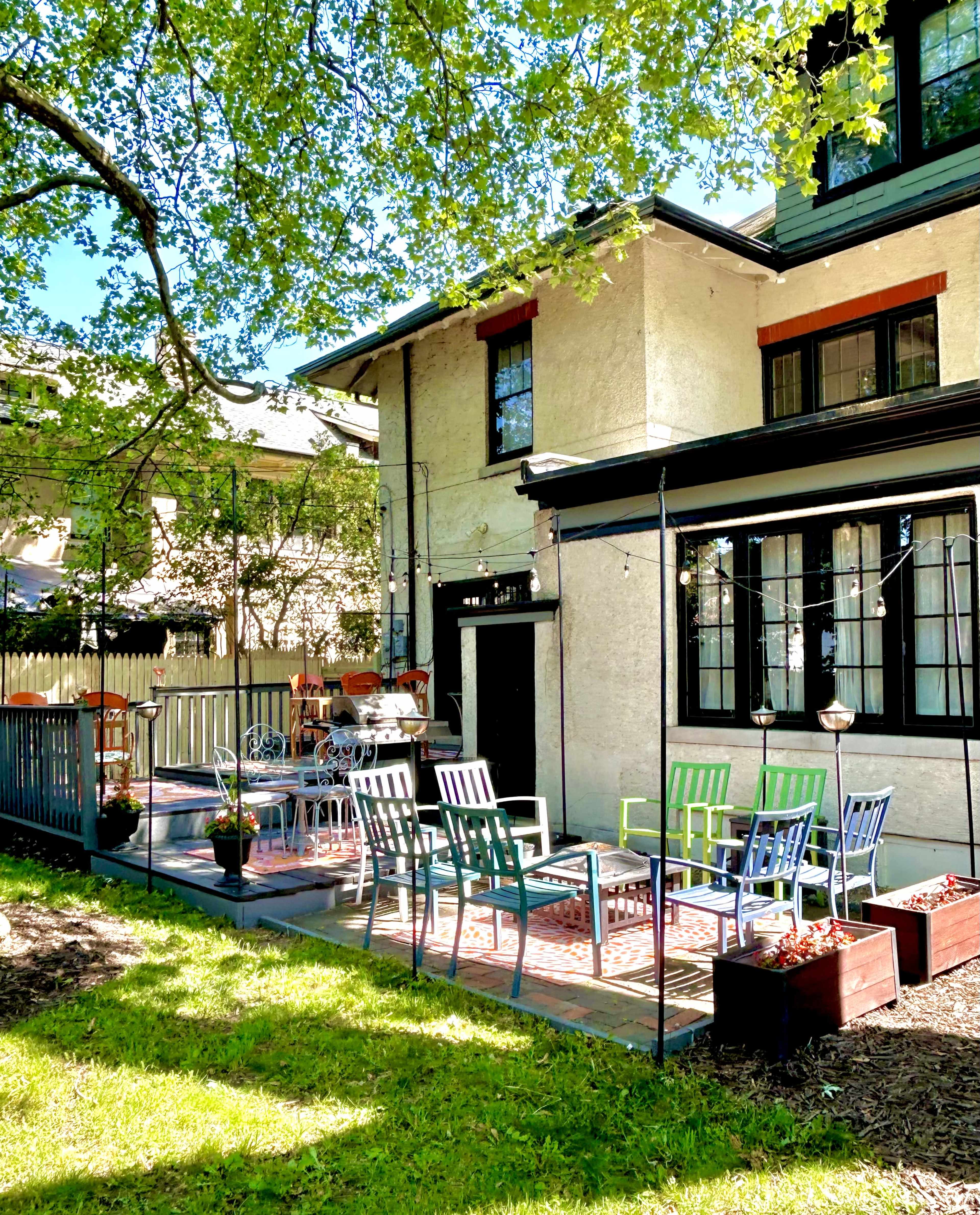 A patio with colorful chairs and tables is set up next to a house, surrounded by greenery and a tree.