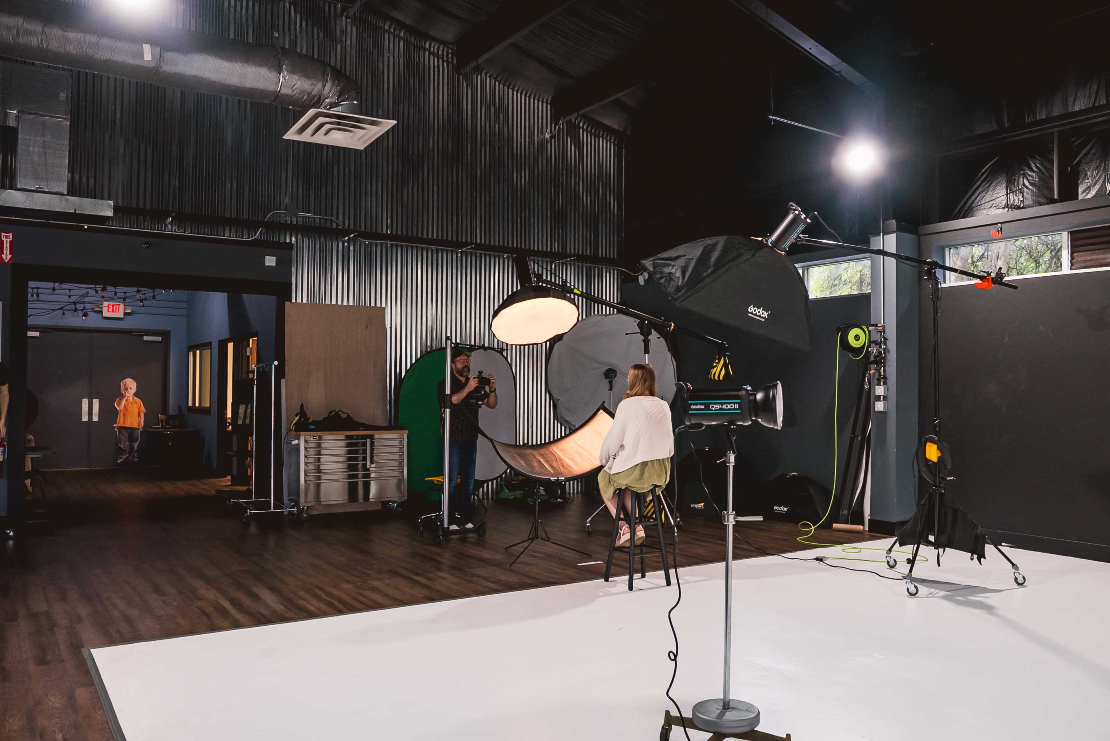 A photography studio is set up with a model seated on a stool, surrounded by lighting equipment and backdrops.