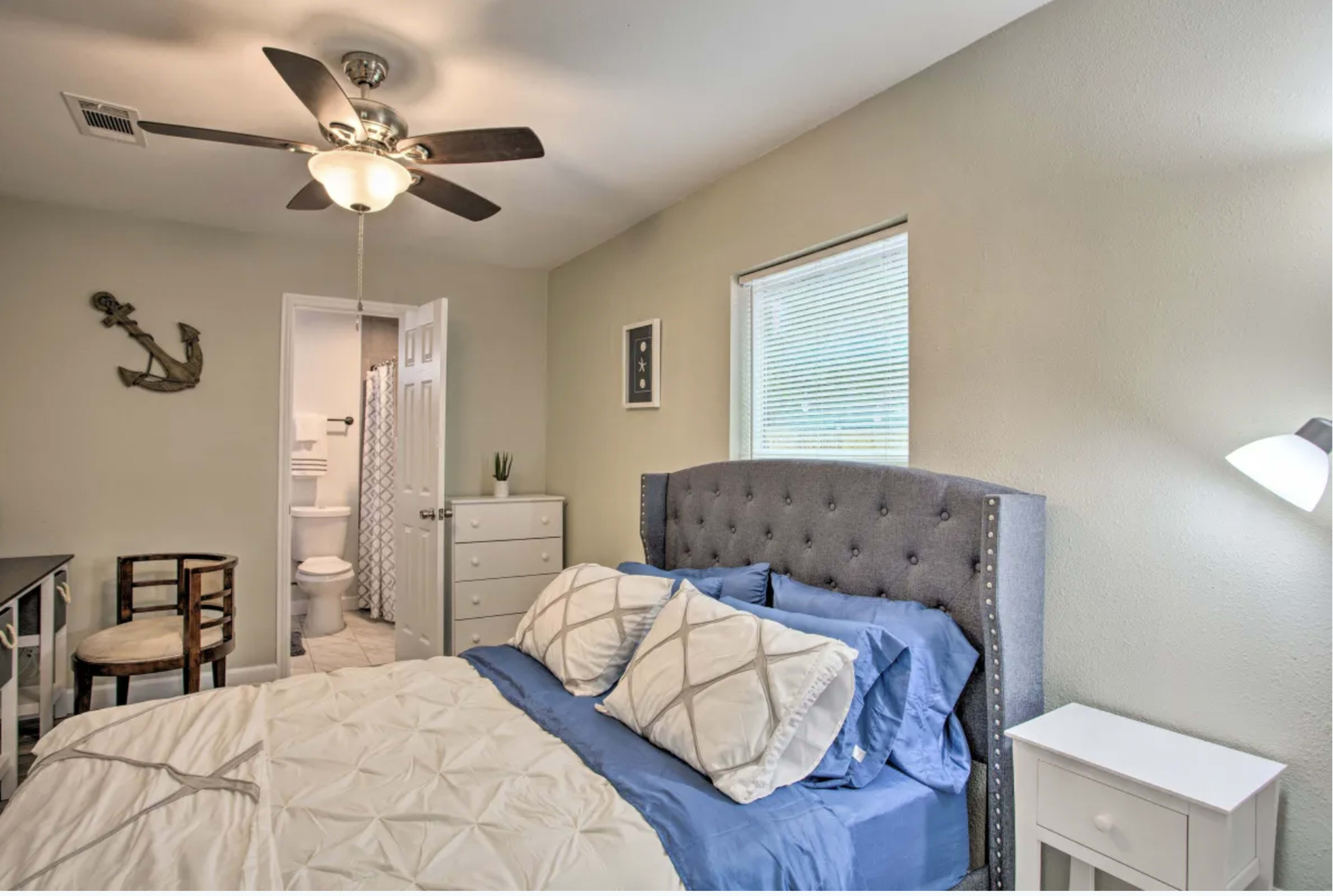 A neatly arranged bedroom with a gray tufted headboard, blue bedding, a small nightstand, and a doorway leading to a bathroom.