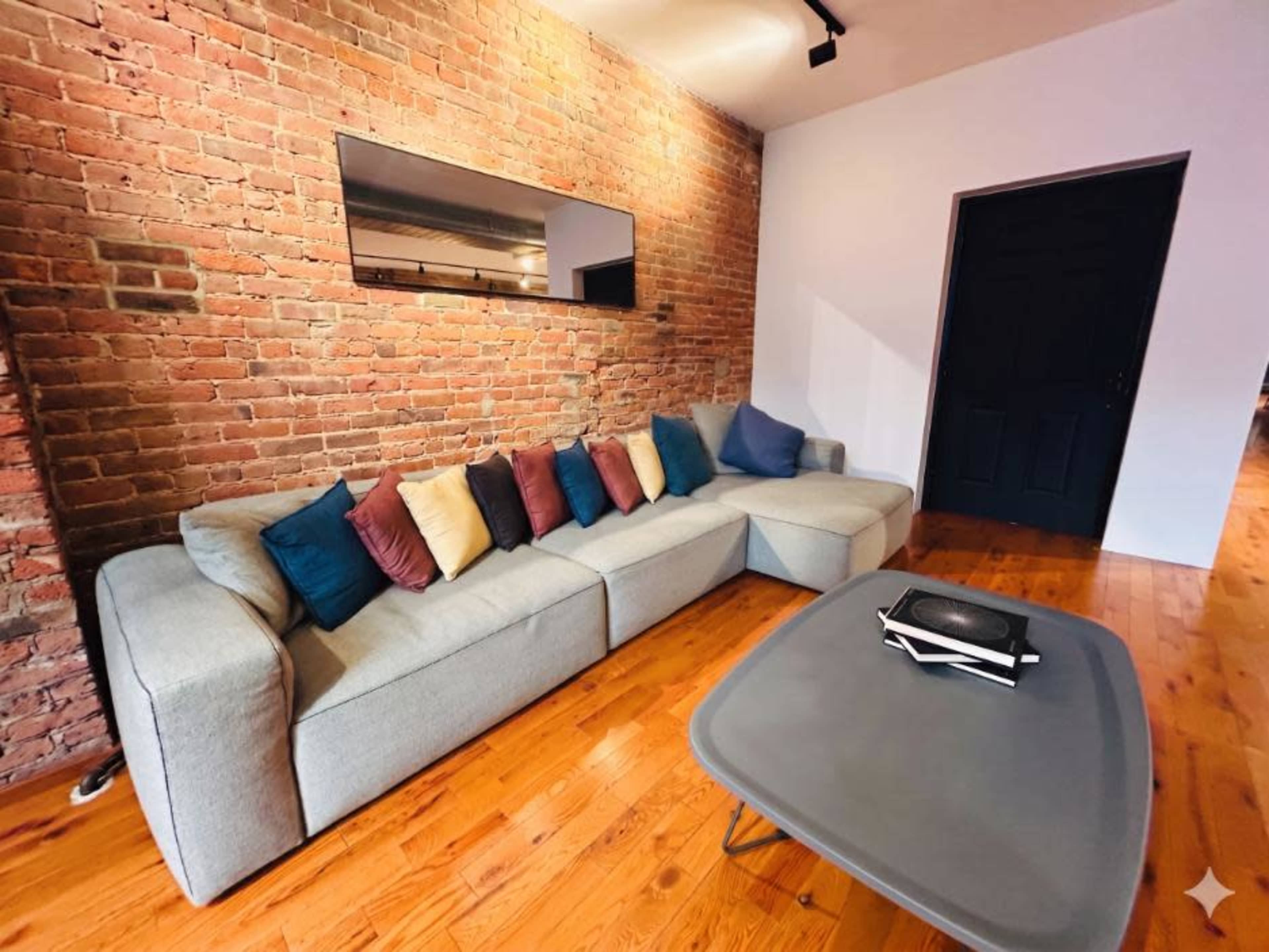 A sectional gray sofa with colorful cushions is positioned in front of a brick wall, alongside a dark wooden table.