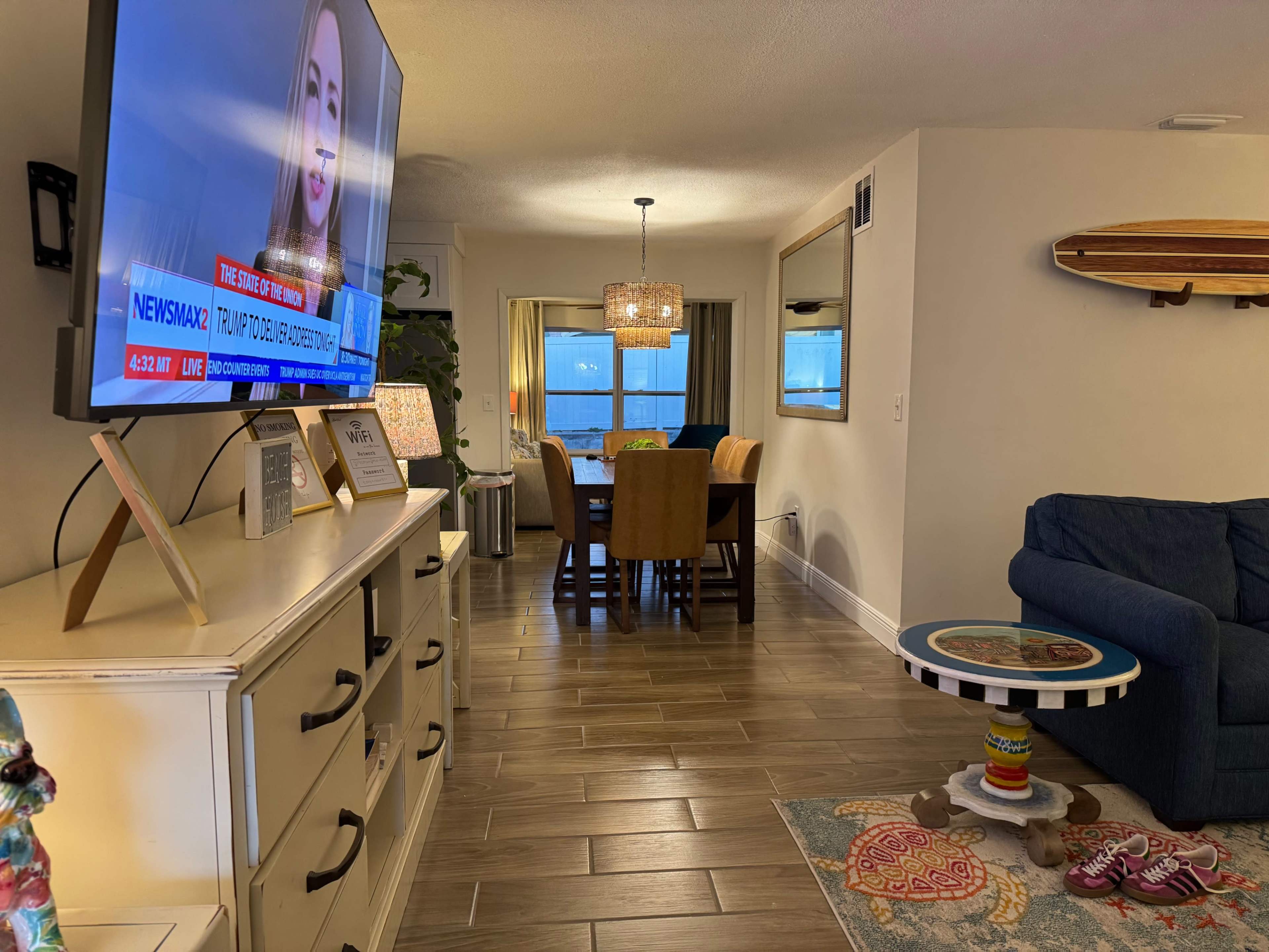 A modern living room features a television displaying news, with a dining area visible in the background and a surfboard mounted on the wall.