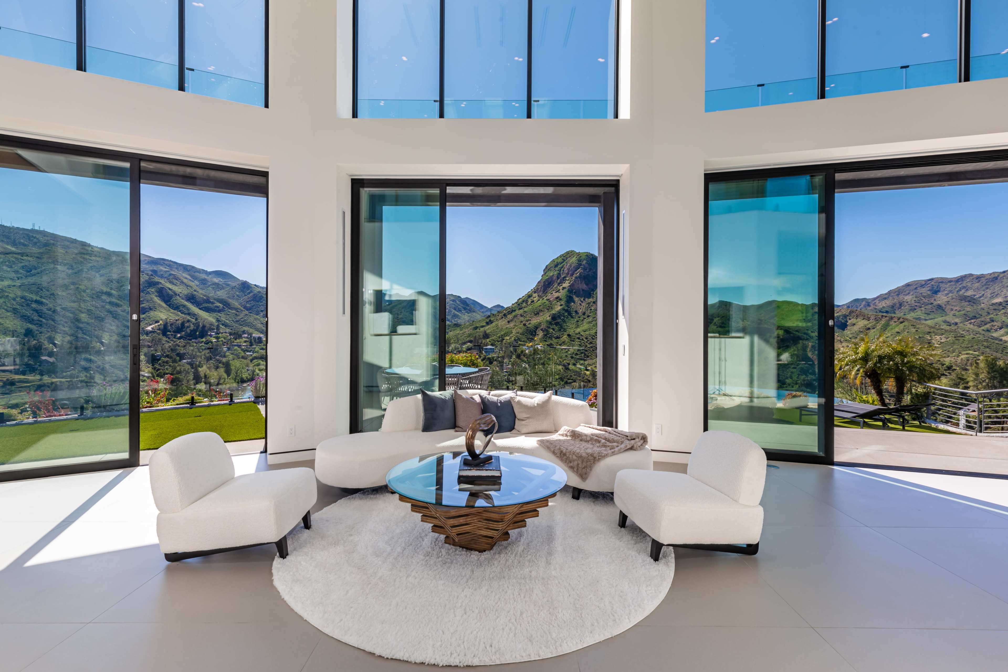 A modern living room features large windows with scenic mountain views, a round glass coffee table, and two white upholstered chairs on a circular rug.
