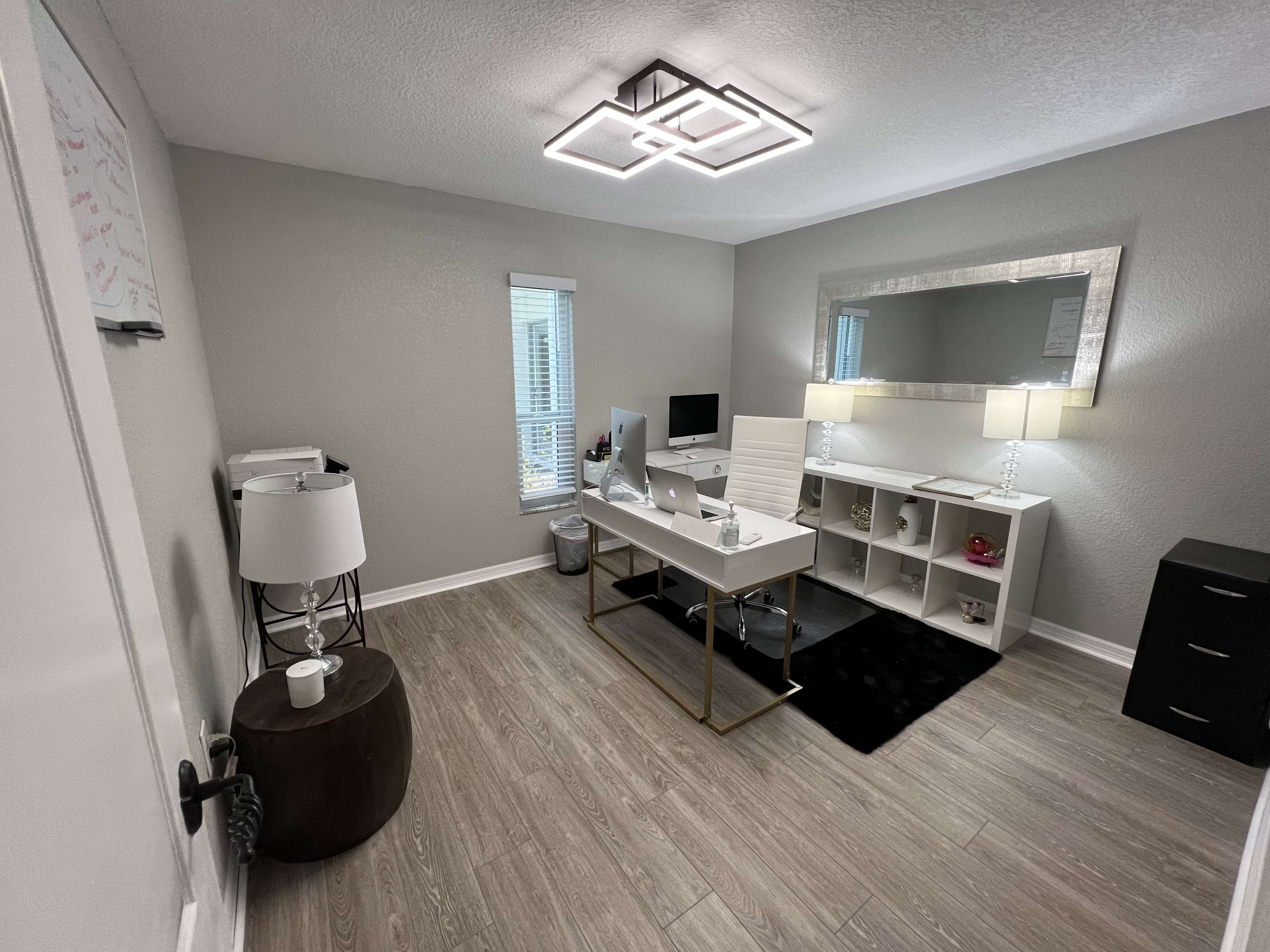 A modern office space featuring a desk with a computer, a large mirror, two lamps, shelving units, and a chair, all arranged on a wood-like floor.