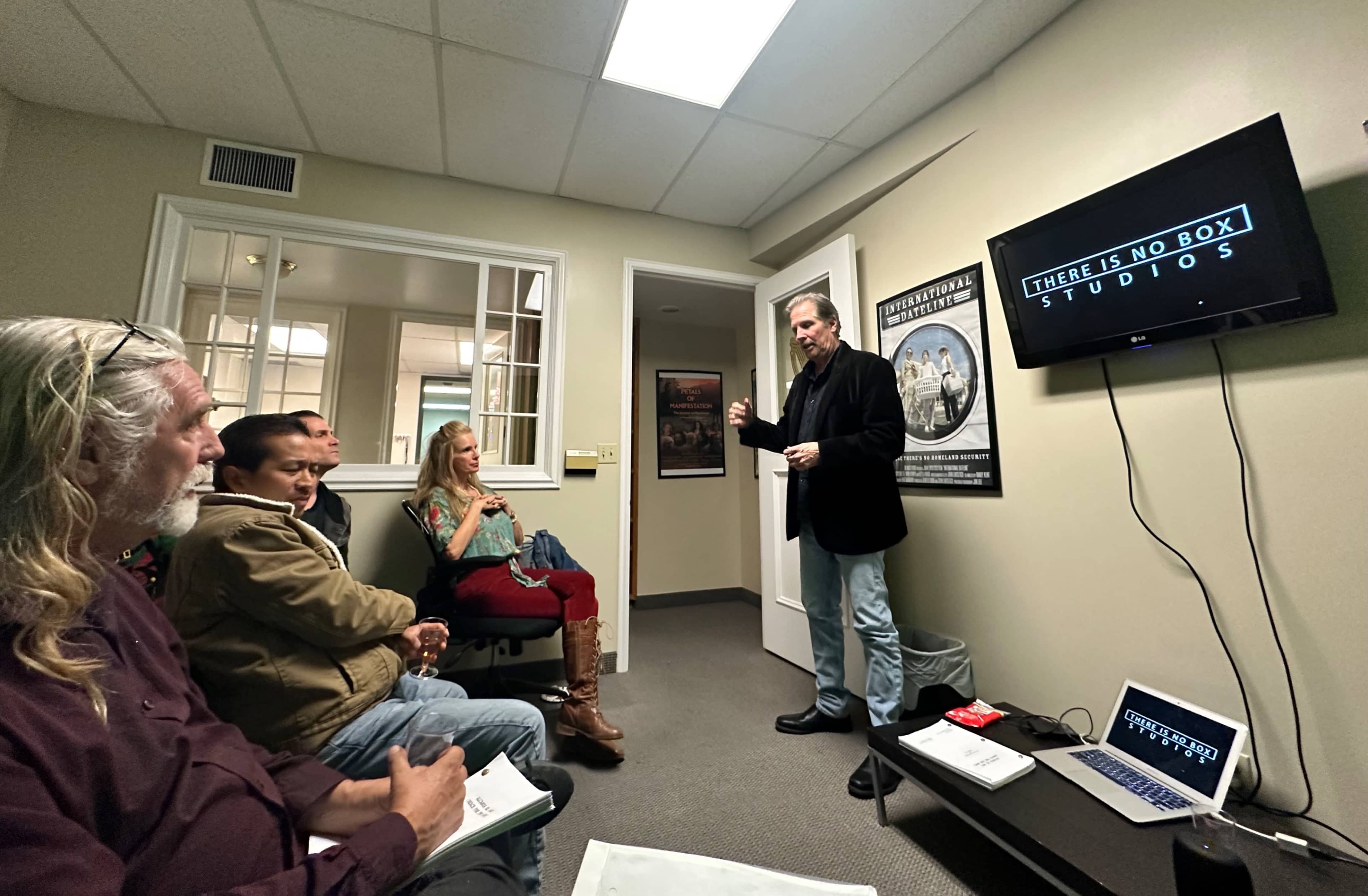 A man stands in front of a screen displaying "THERE IS NO BOX STUDIOS," while a small audience of three sits attentively in a room with light-colored walls and framed posters.