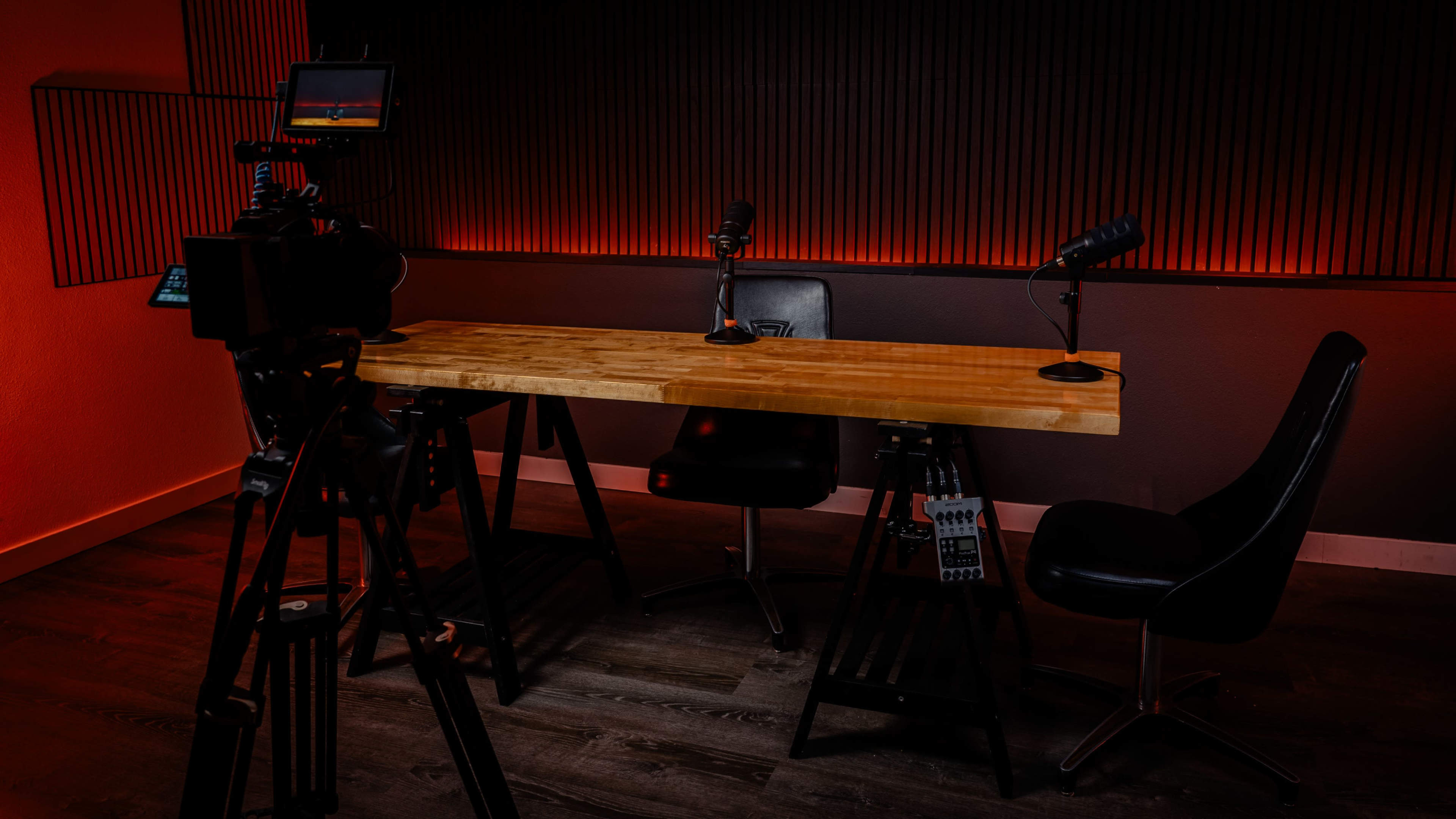 A camera is set up in front of a wooden table with two chairs and microphones, all illuminated by red lighting against a dark wall.