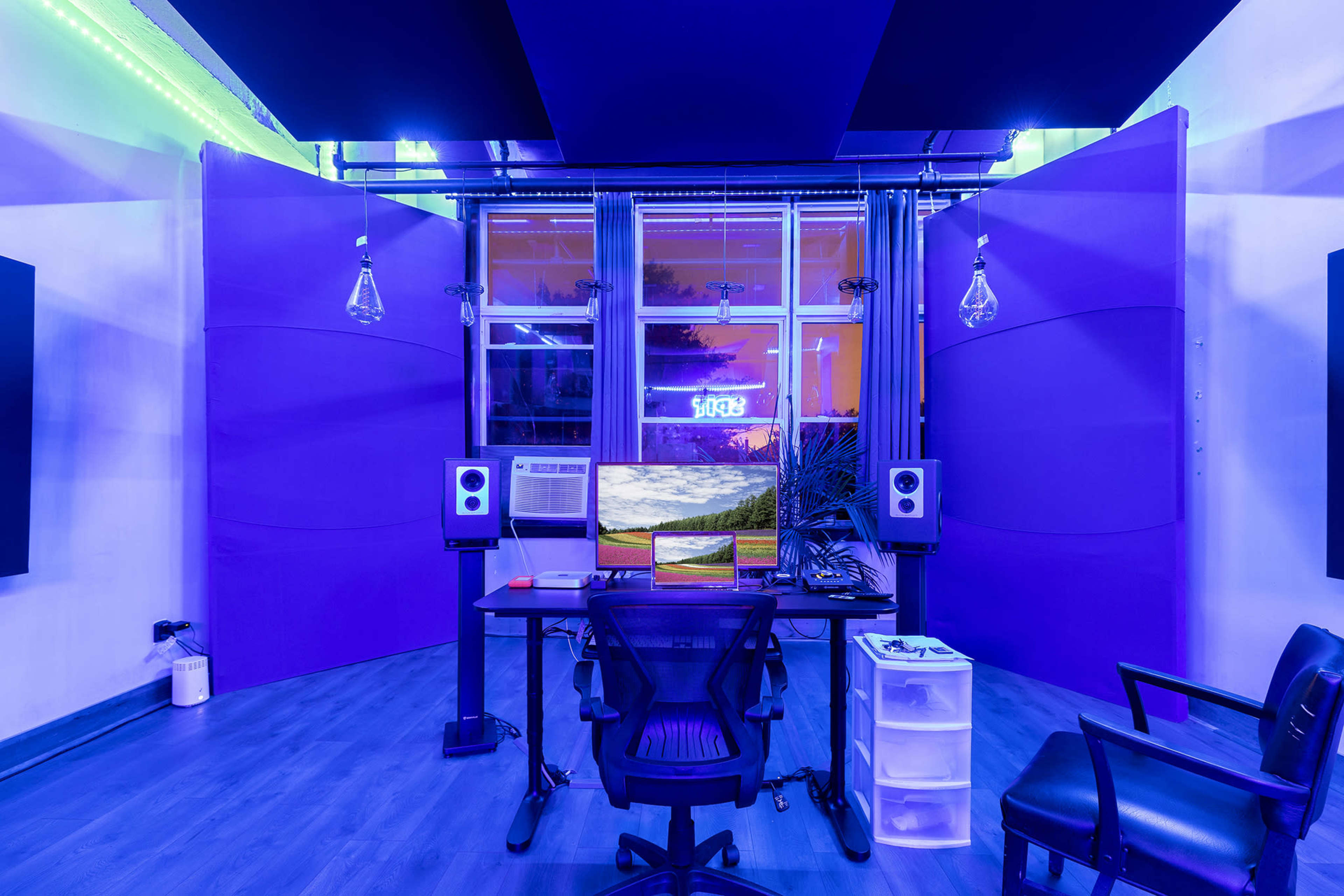 A modern music studio featuring a desk with monitors, studio speakers, and purple acoustic panels against a backdrop of illuminated windows.