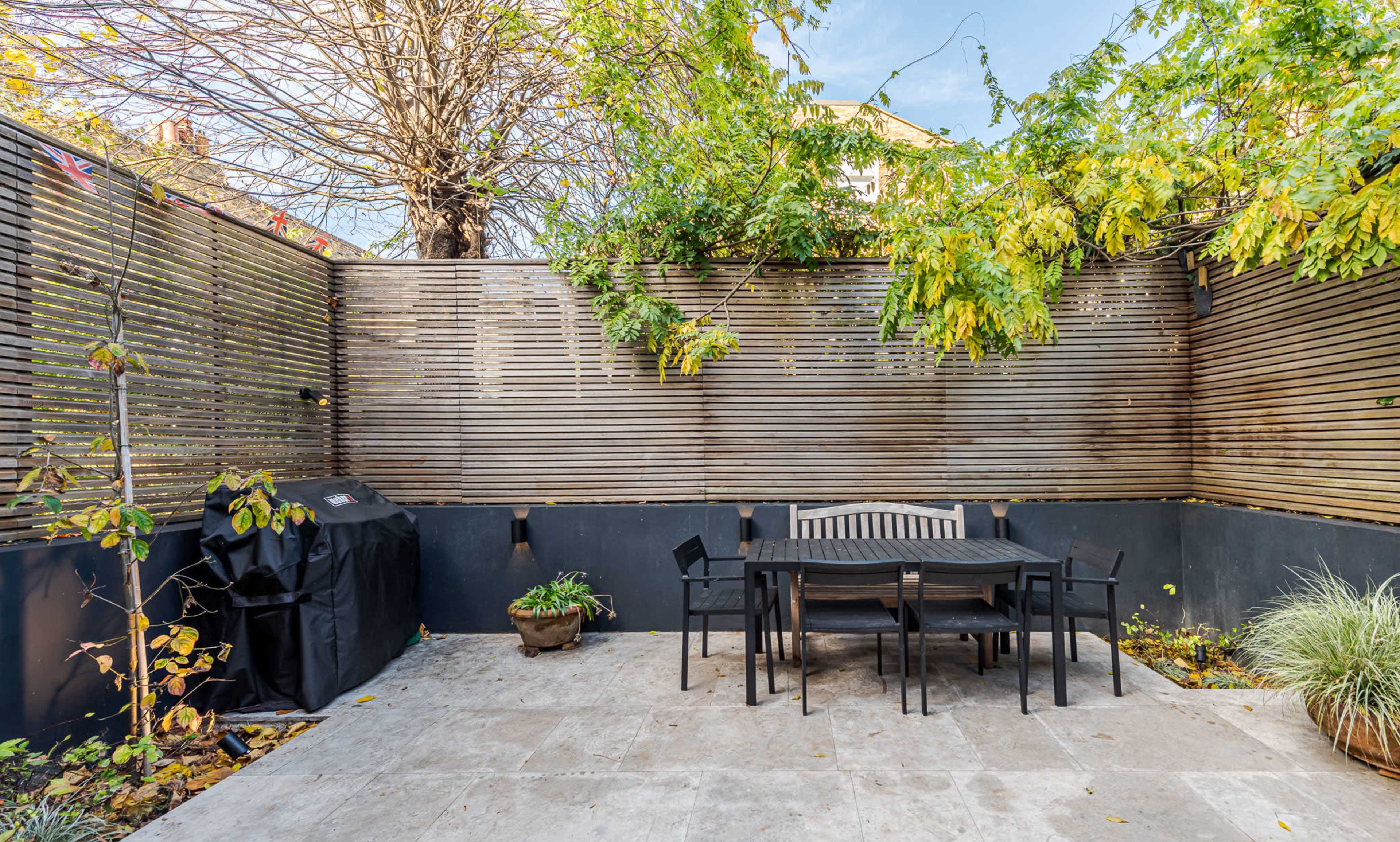 The image shows a small outdoor patio with a wooden dining table and chairs, surrounded by wooden fencing and some greenery.