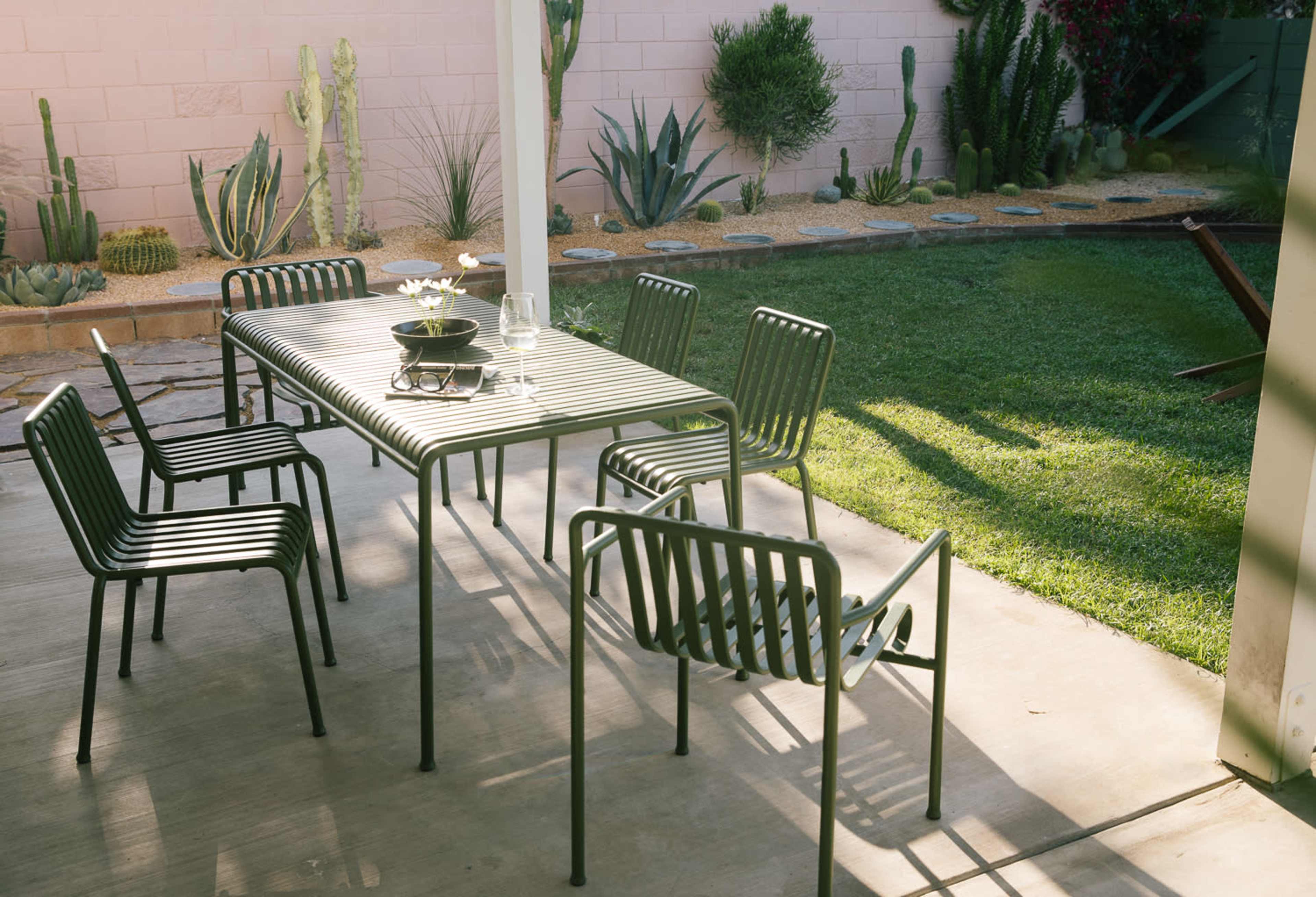 A metal dining table surrounded by six chairs is positioned on a patio with a vibrant garden featuring cacti and grass in the background.