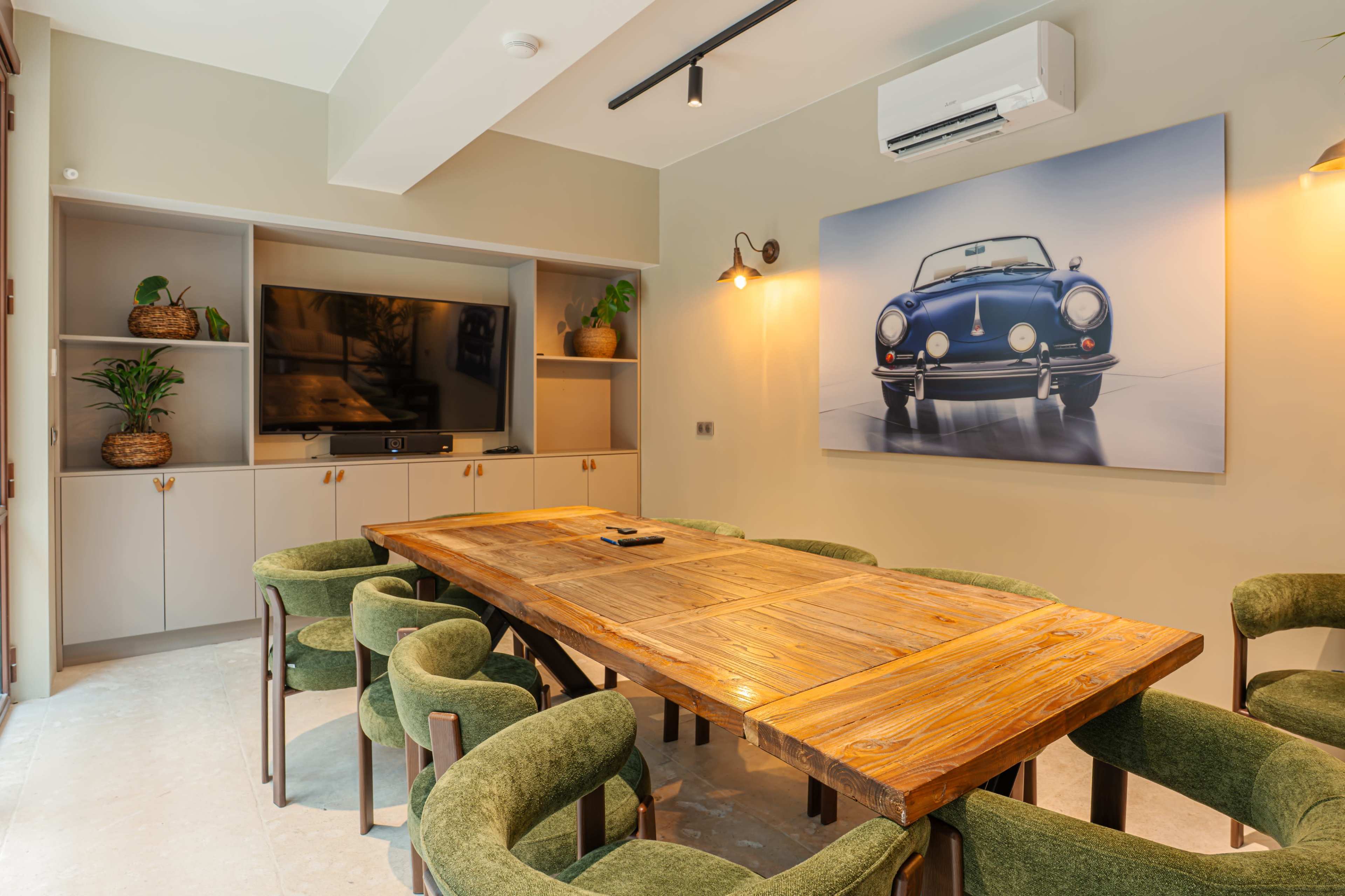The image shows a modern meeting room featuring a large wooden table surrounded by green upholstered chairs, with a television and a car artwork on the wall.