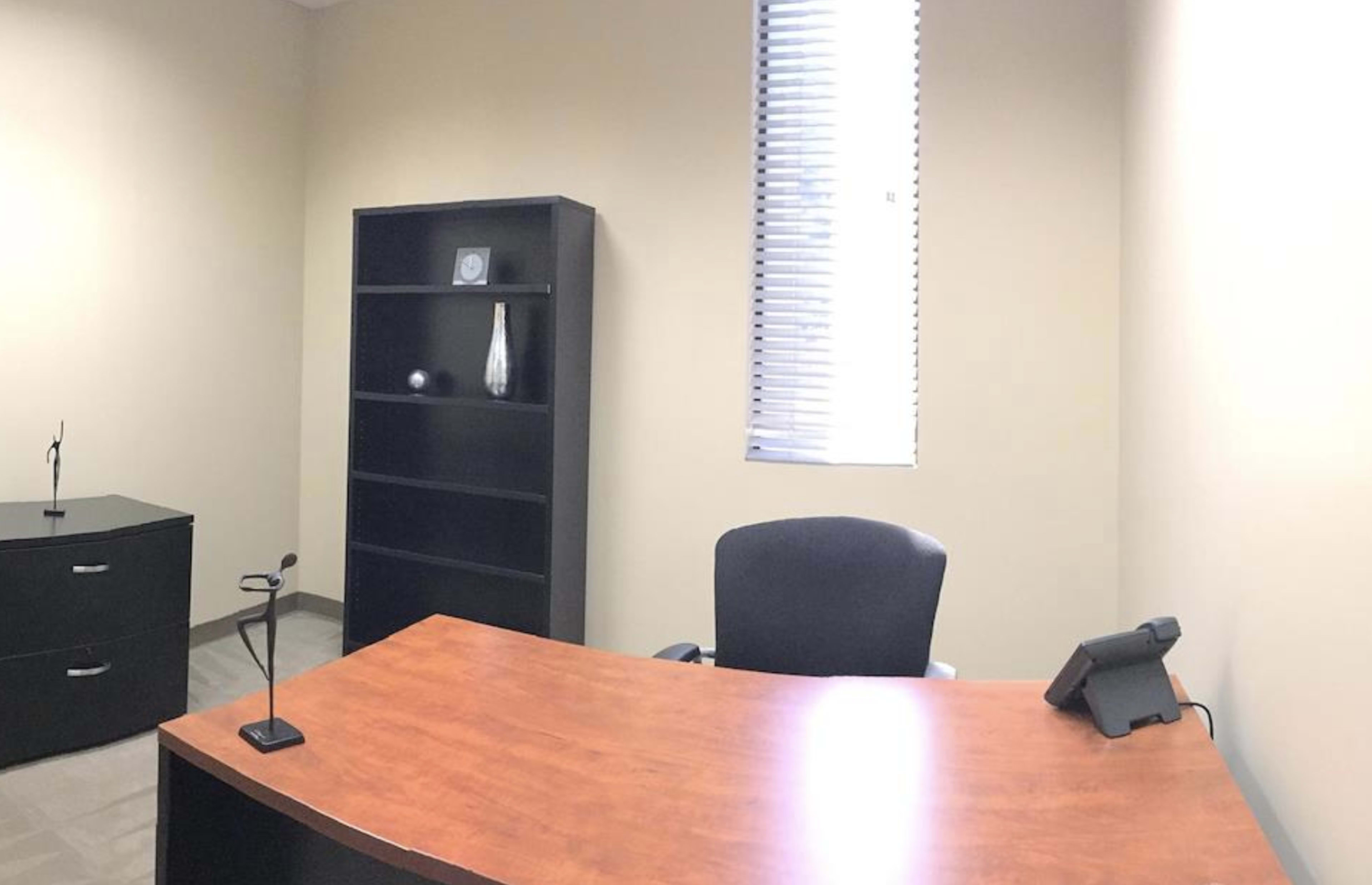 The image shows an office space with a wooden desk, a chair, a black filing cabinet, and a bookshelf against a plain wall.