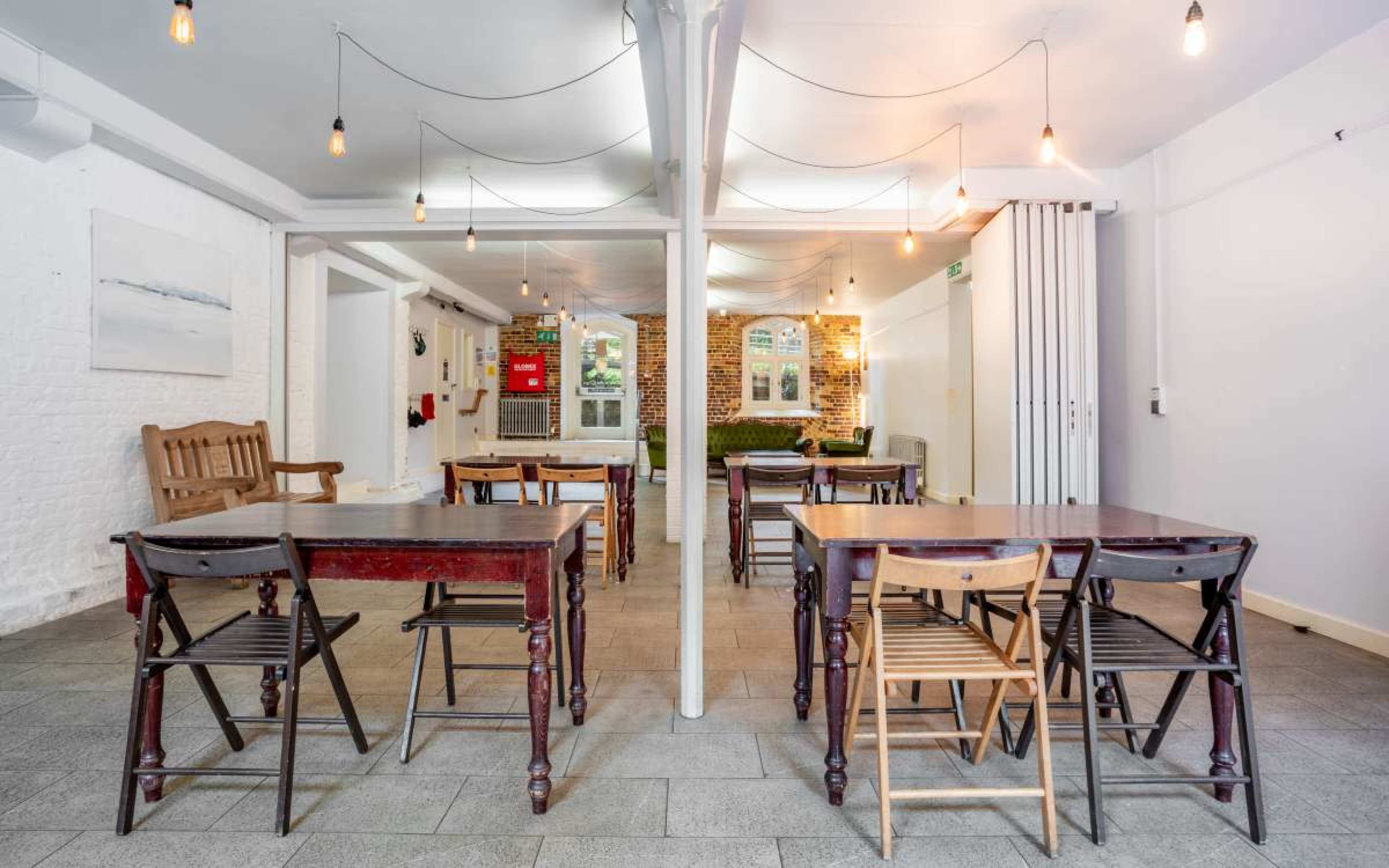 A spacious, well-lit room with several wooden tables and chairs arranged for seating, featuring exposed brick walls and hanging light fixtures.