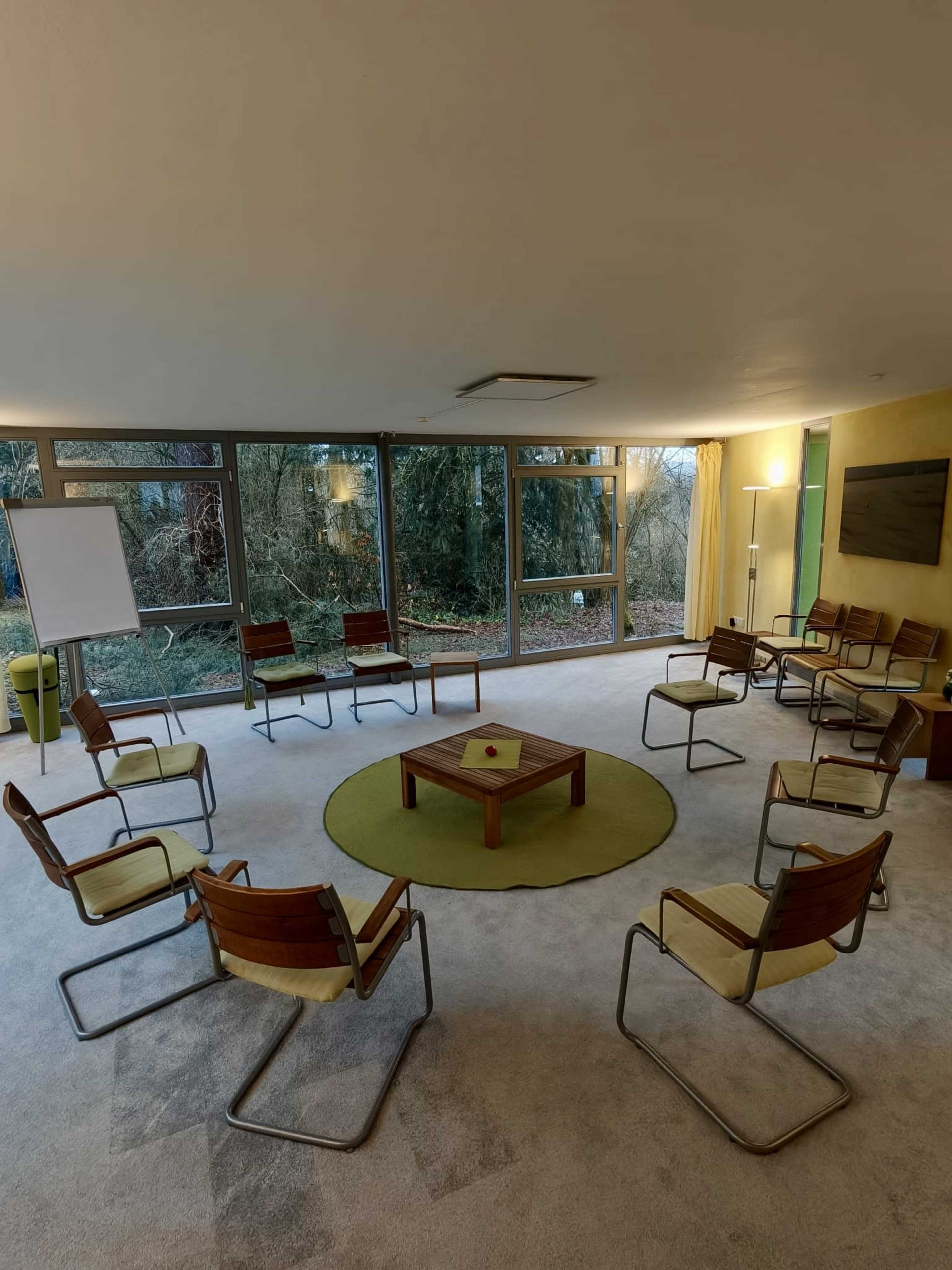The image shows a conference room with a circular arrangement of eight chairs around a wooden table, situated in front of large windows that overlook a wooded area.