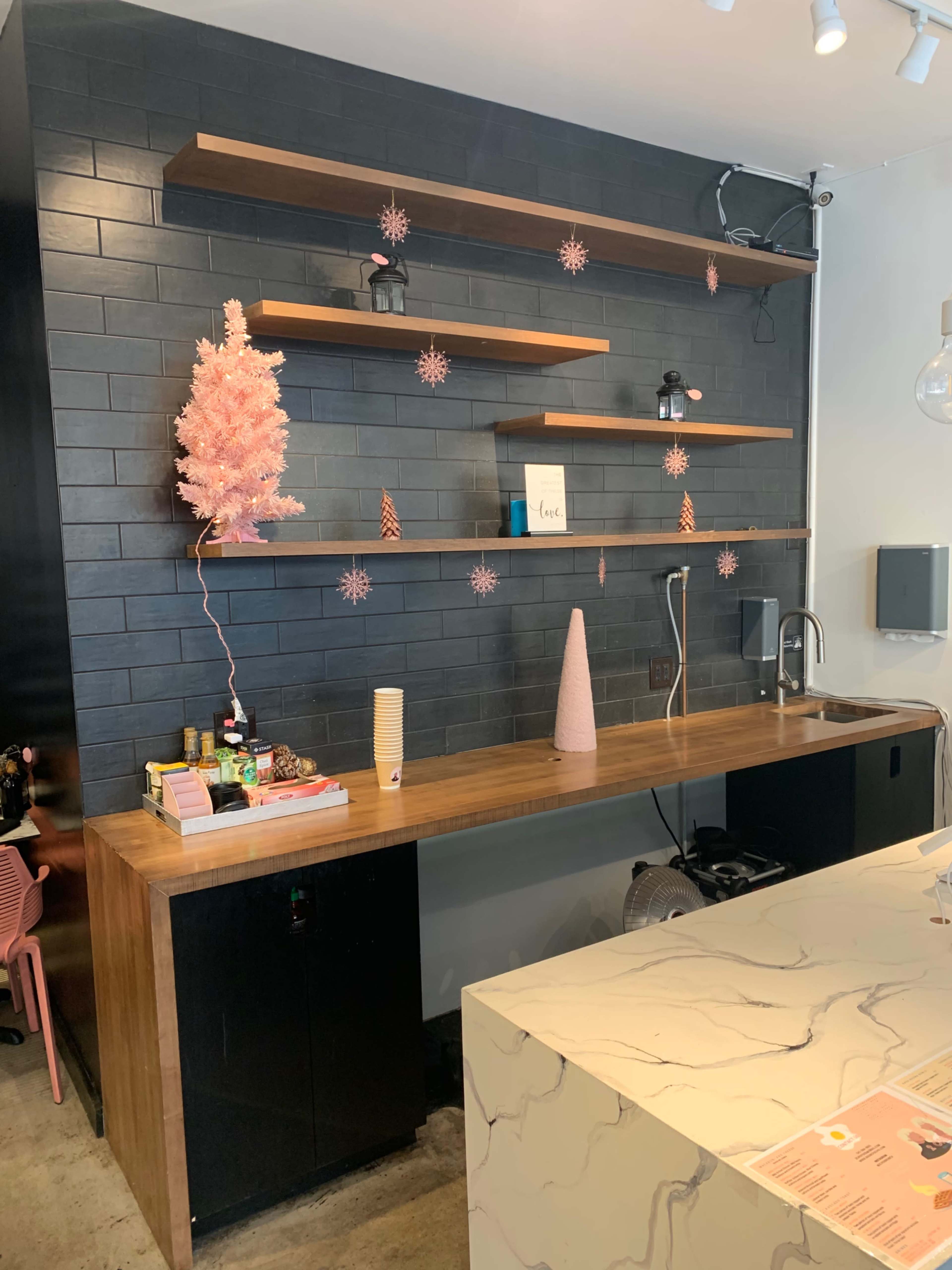 A minimalist café interior featuring a dark tiled wall, wooden shelves displaying decorative items, and a countertop with various supplies and a pink Christmas tree.
