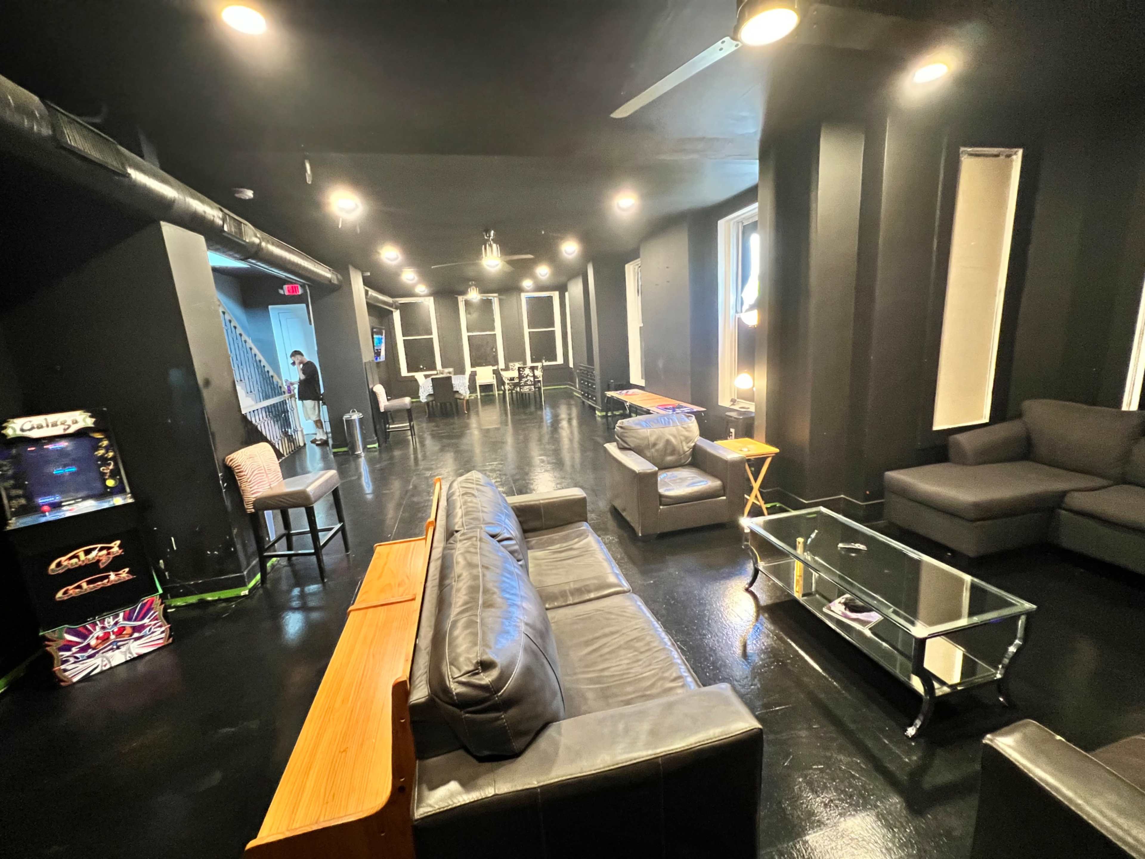 A spacious lounge area furnished with sofas, a glass coffee table, and an arcade machine, featuring black walls and flooring.