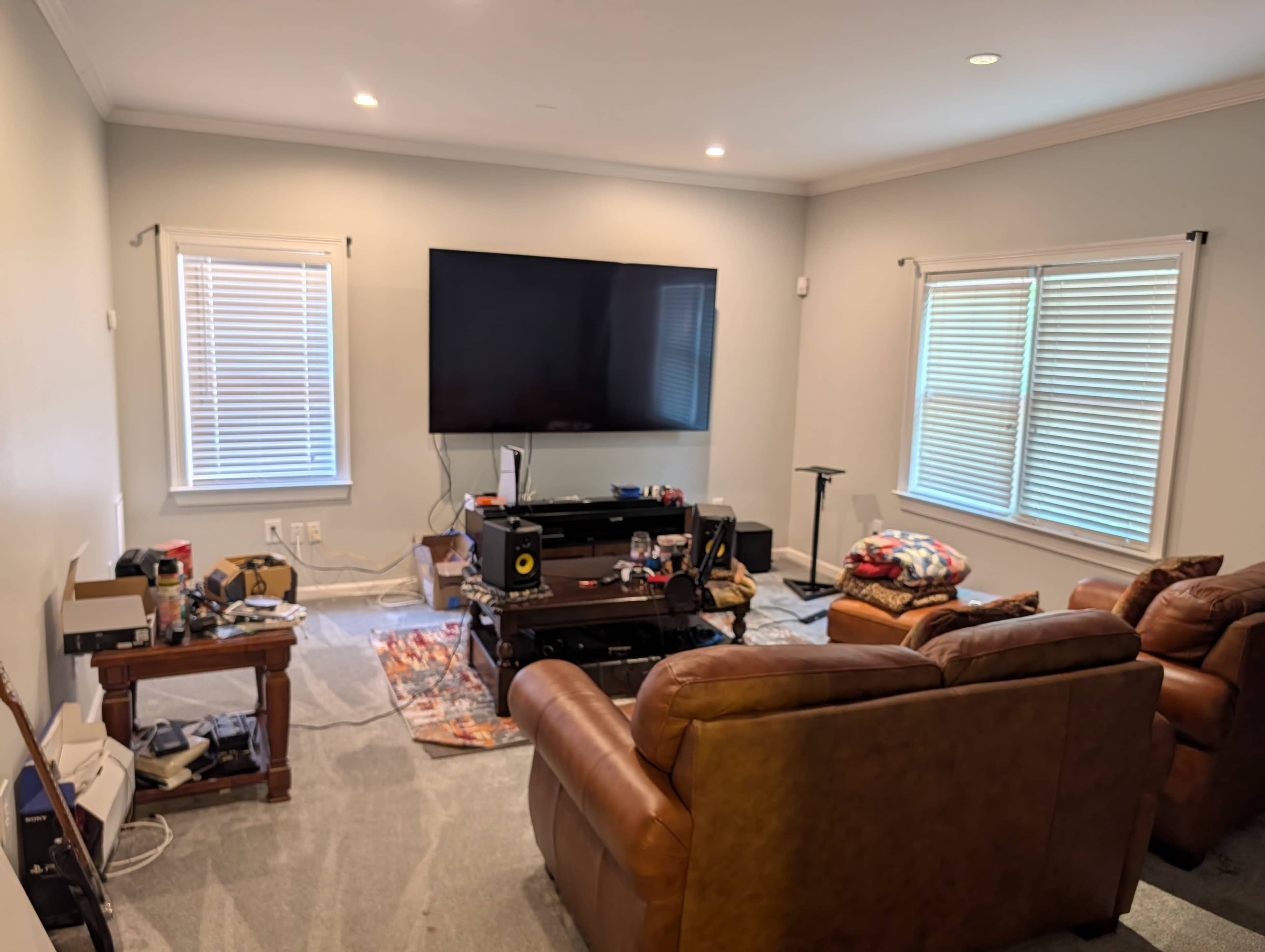 The image shows a living room with a large wall-mounted TV, a brown leather sofa, and various items scattered around, including furniture and a rug on the carpeted floor.