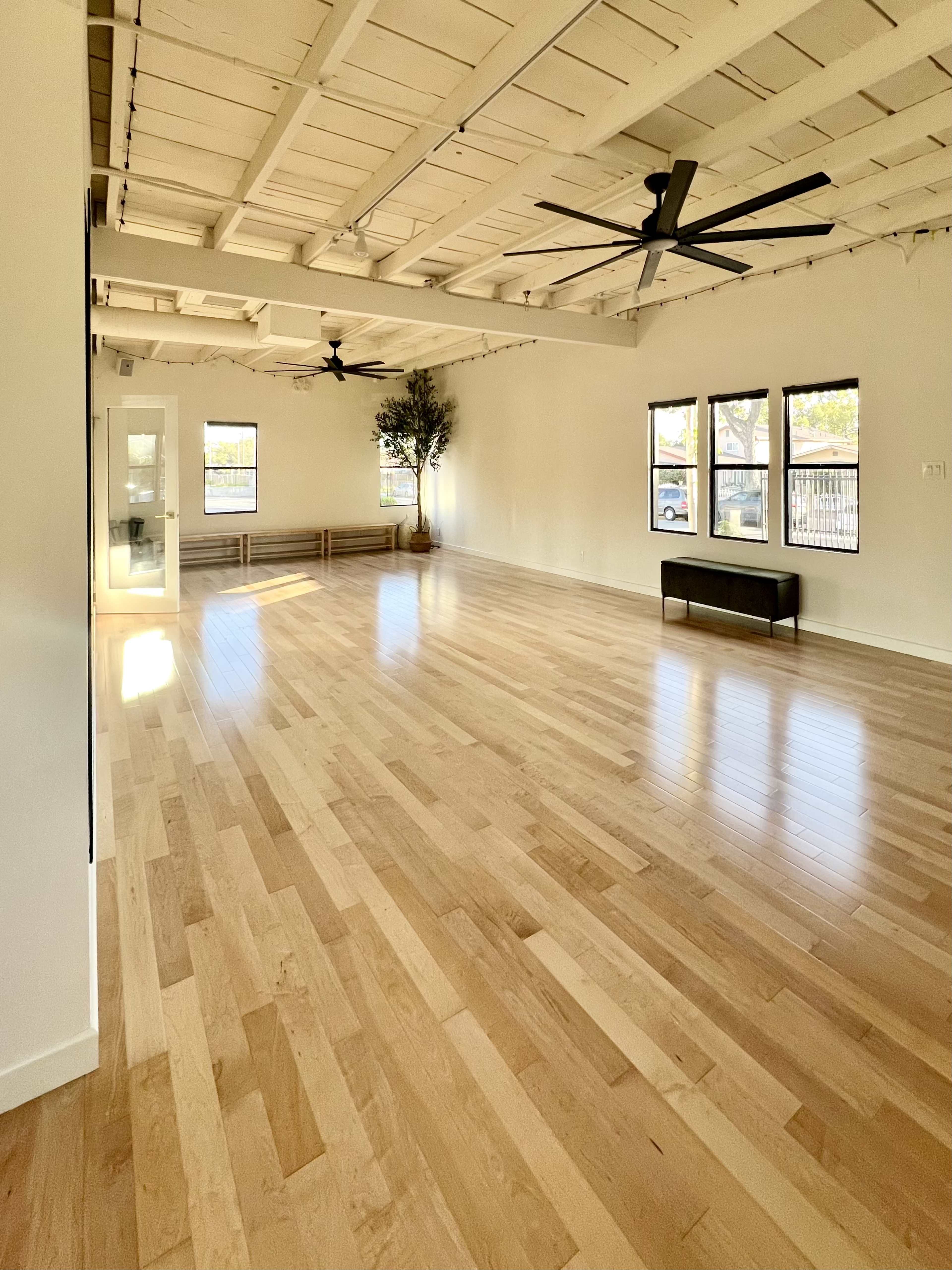 A spacious, well-lit room with wooden flooring, two ceiling fans, and large windows.