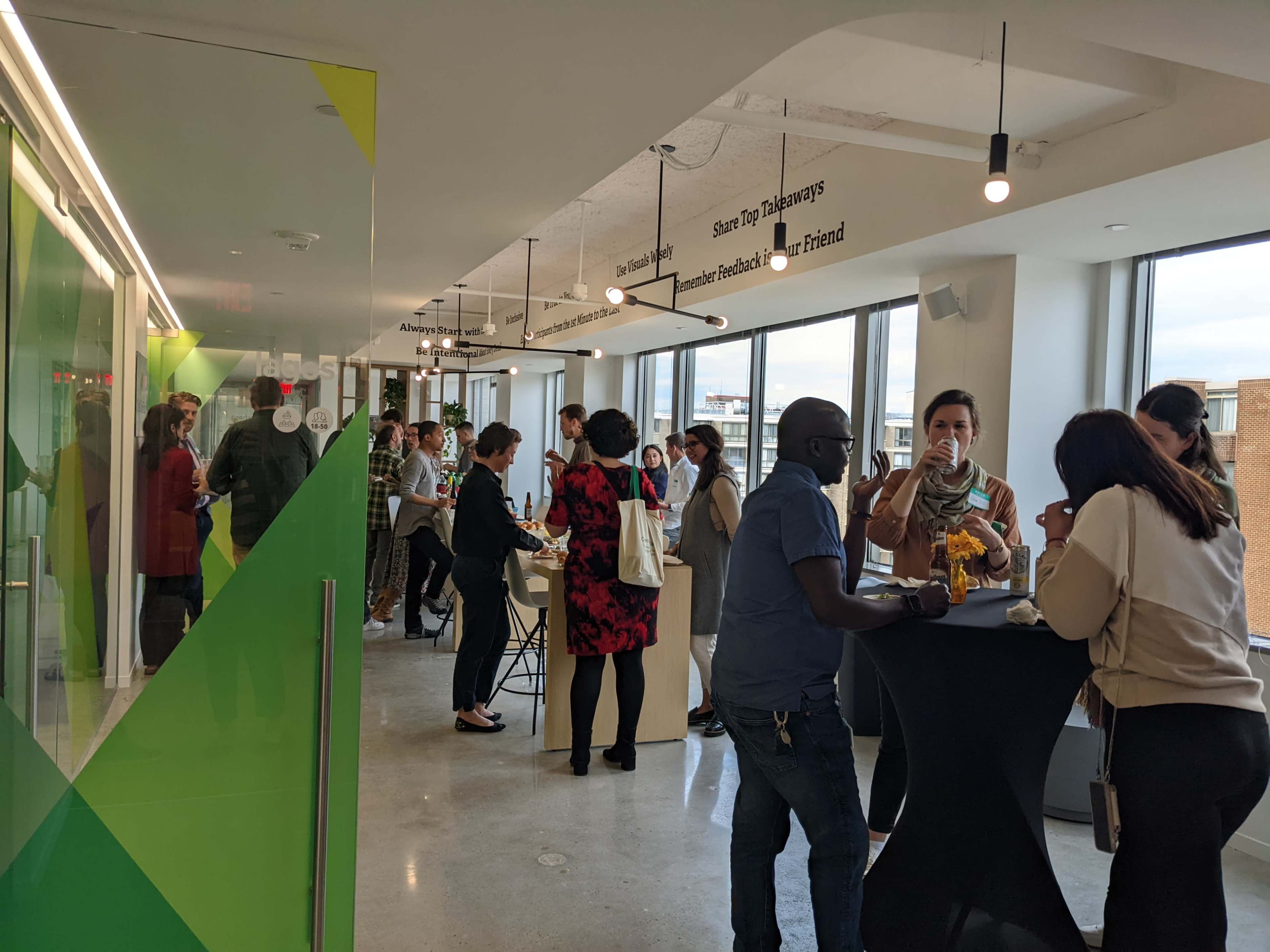A group of people mingles and converses at a networking event in a brightly lit office space with large windows.