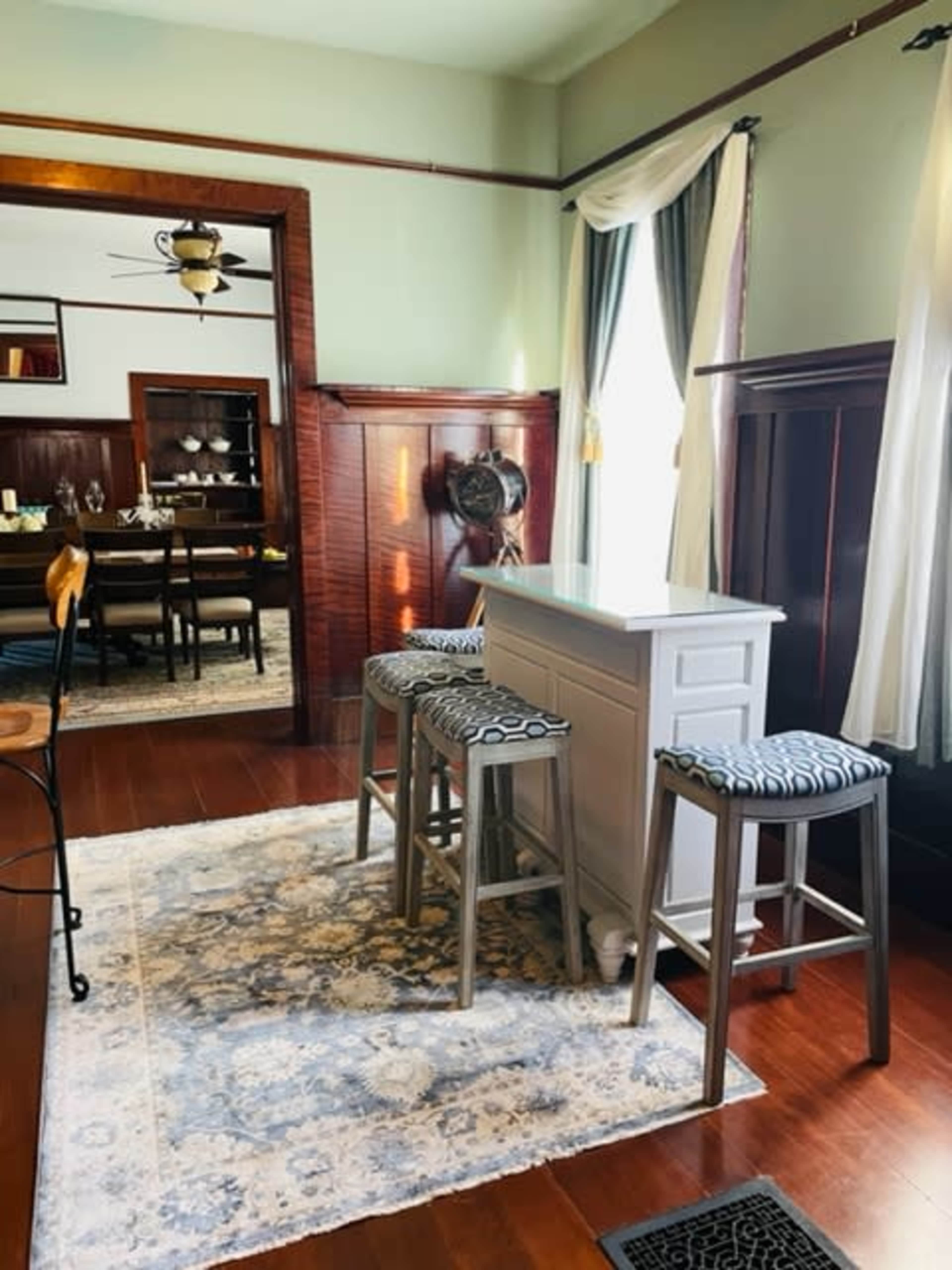 A cozy dining area featuring a small bar with stools and a dining table visible in the background, all set within a room with hardwood floors and large windows.