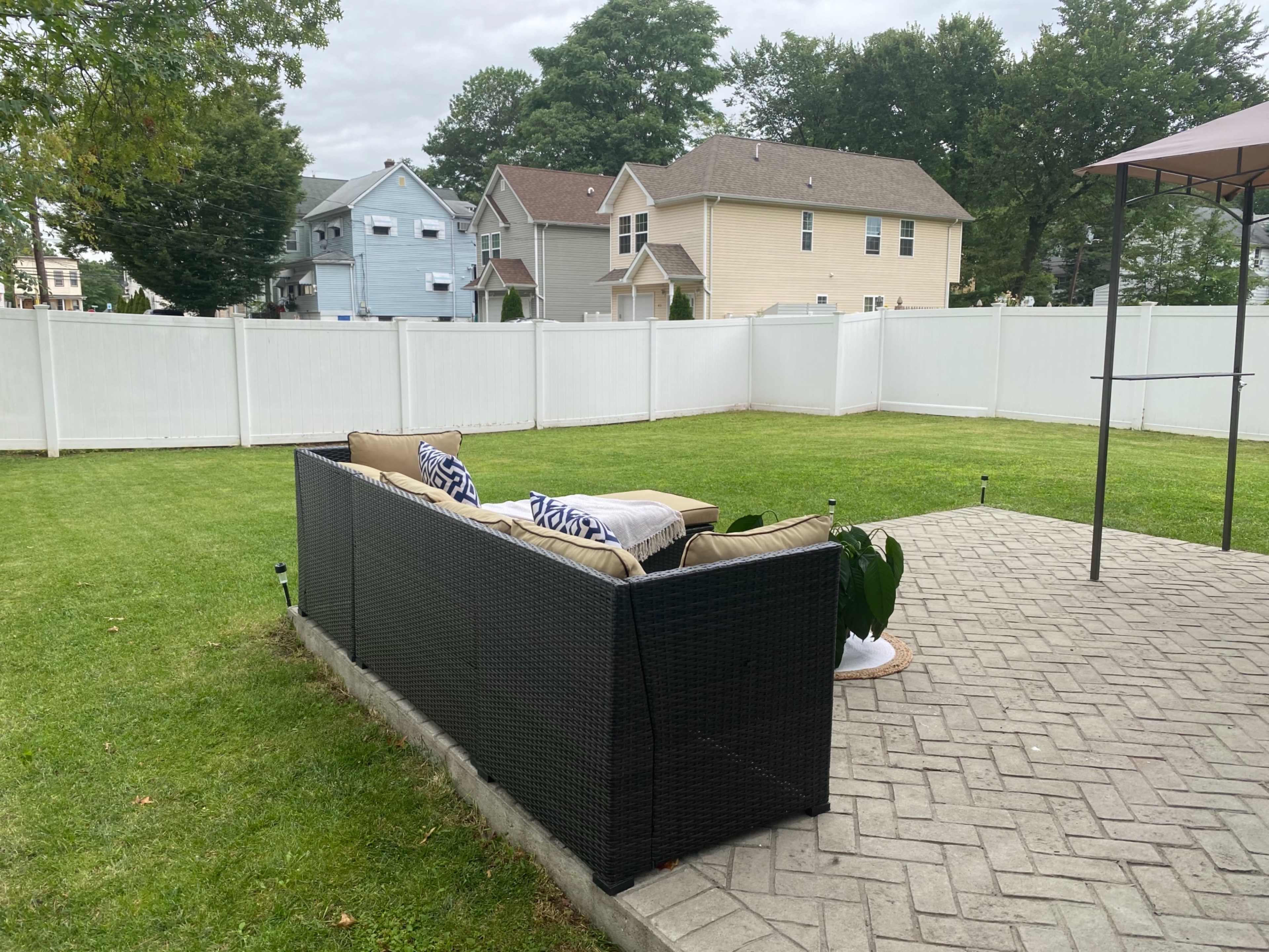 A backyard features a patio with a black wicker sofa, surrounded by a white fence and green lawn.