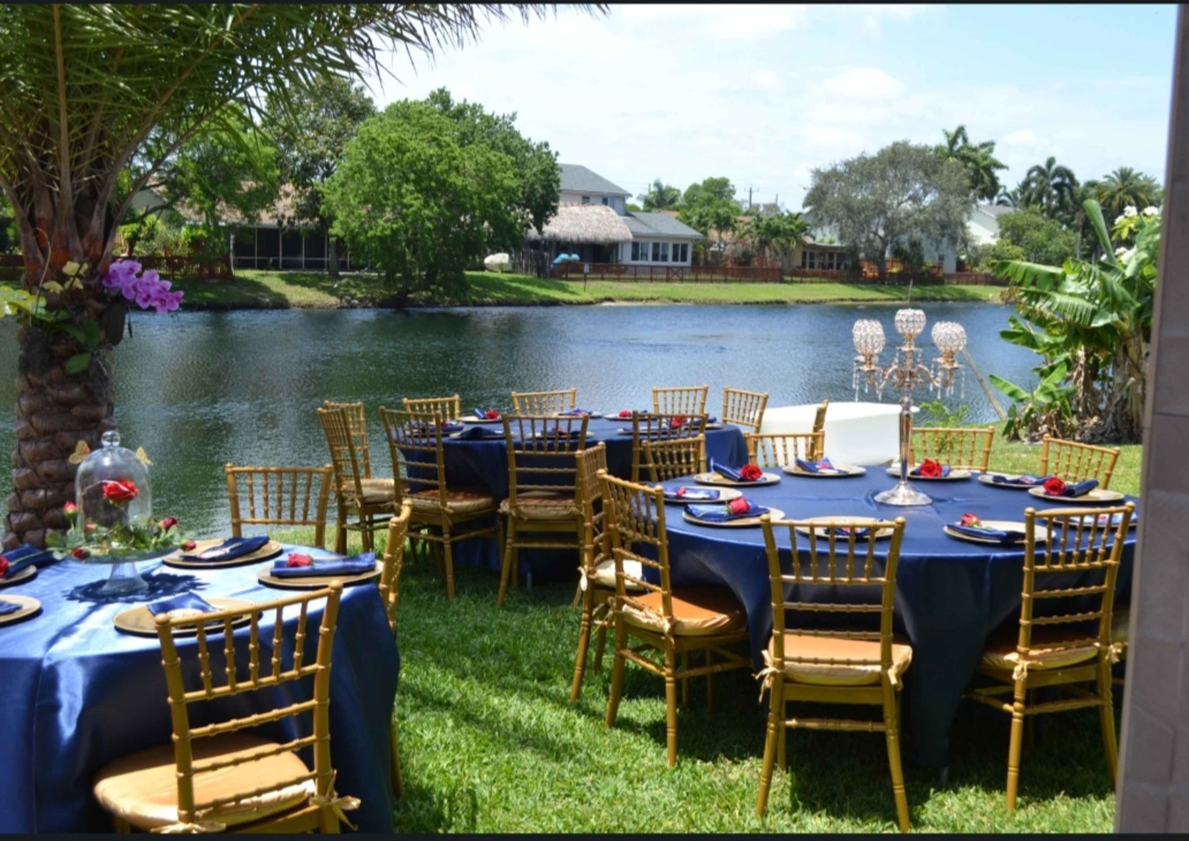 A beautifully arranged outdoor dining setup with golden chairs and blue tablecloths overlooks a serene pond.