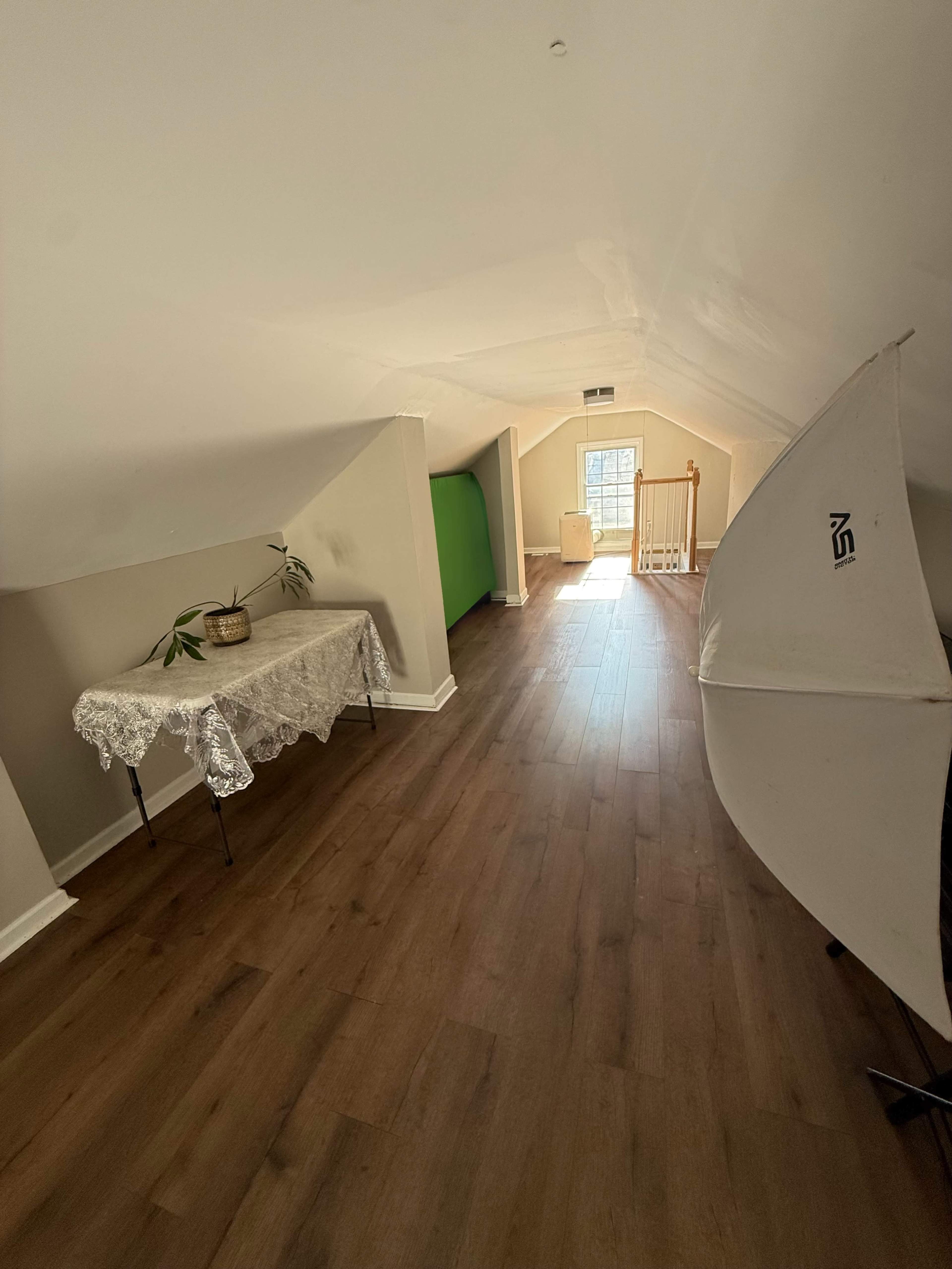 The image shows a well-lit attic space with wooden flooring, a table covered with a lace cloth, a green backdrop, and a white reflector setup.