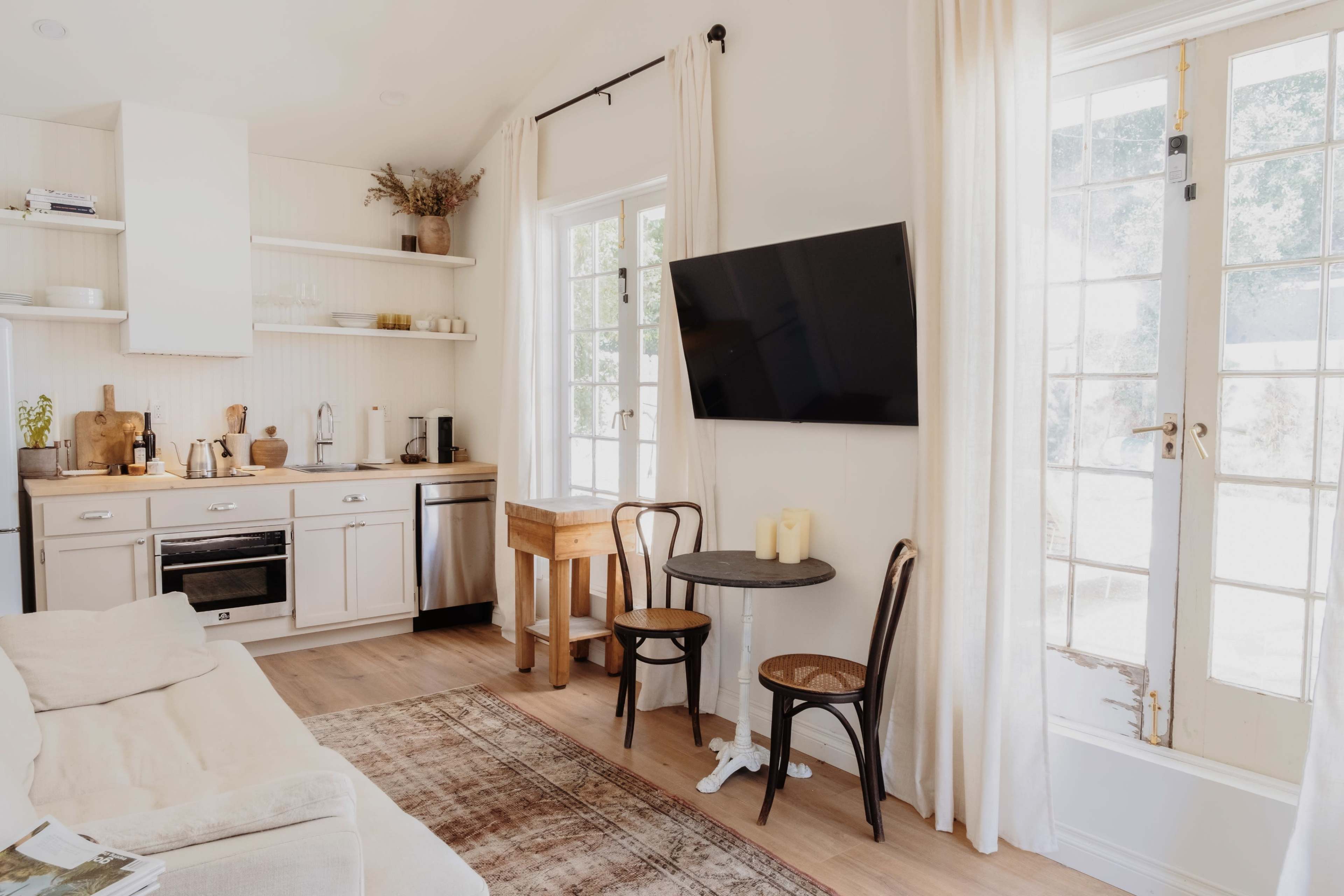 The image shows a cozy, modern interior with a kitchen area, a small dining table with two chairs, and a TV mounted on the wall beside large windows.