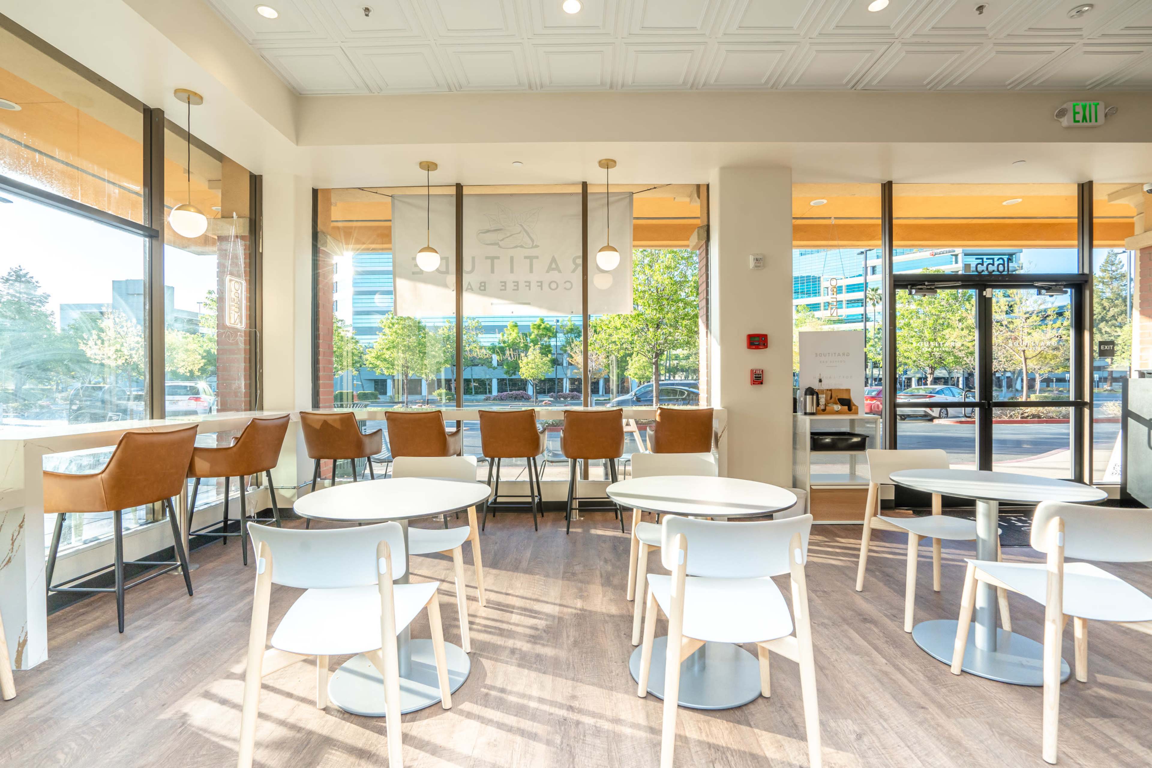 A modern coffee shop interior with large windows, light wooden flooring, and minimalist furniture, including round tables and bar stools.