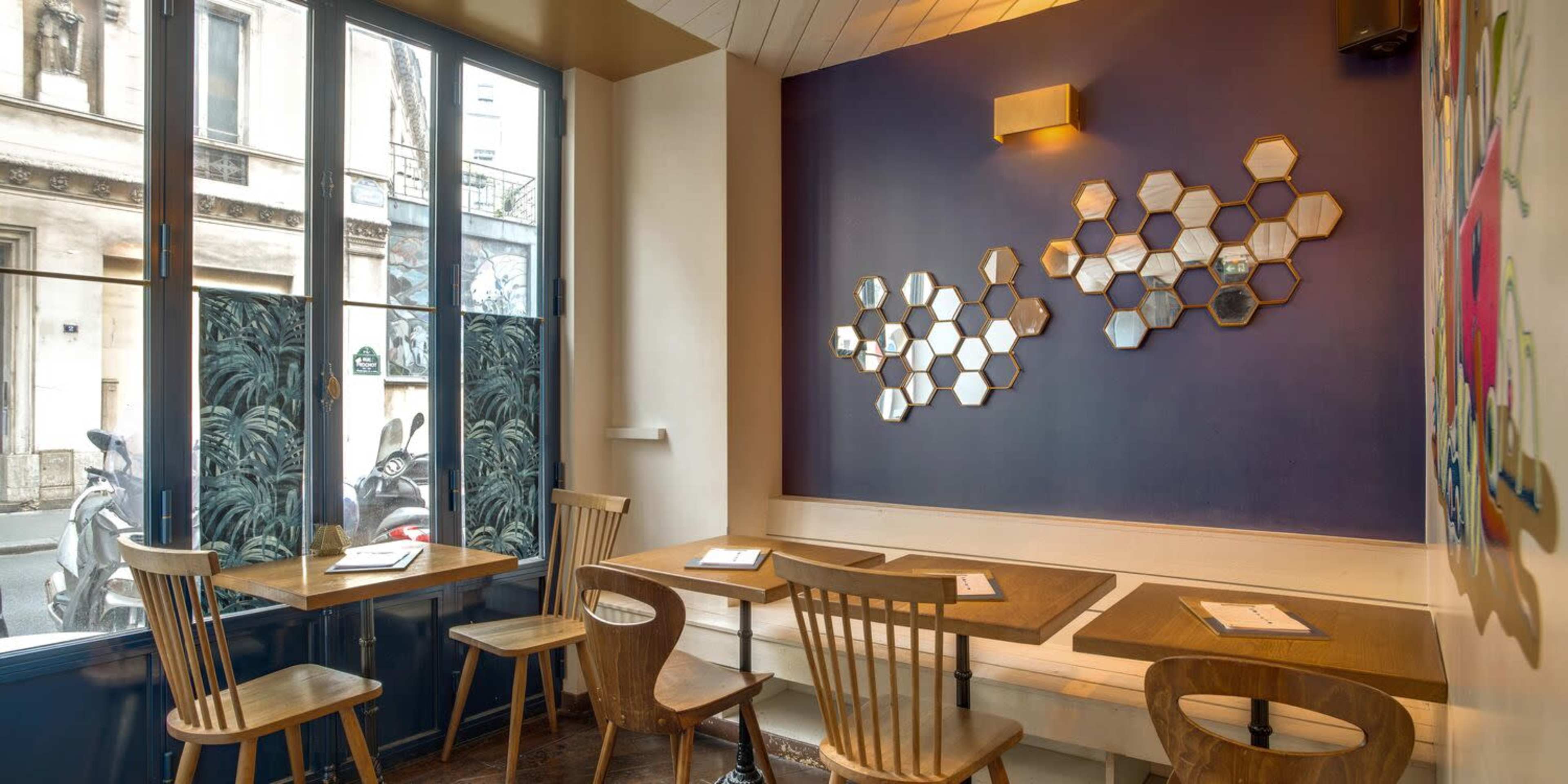 The image shows a café interior with wooden tables and chairs, a wall adorned with hexagonal mirrors, and large windows facing the street.