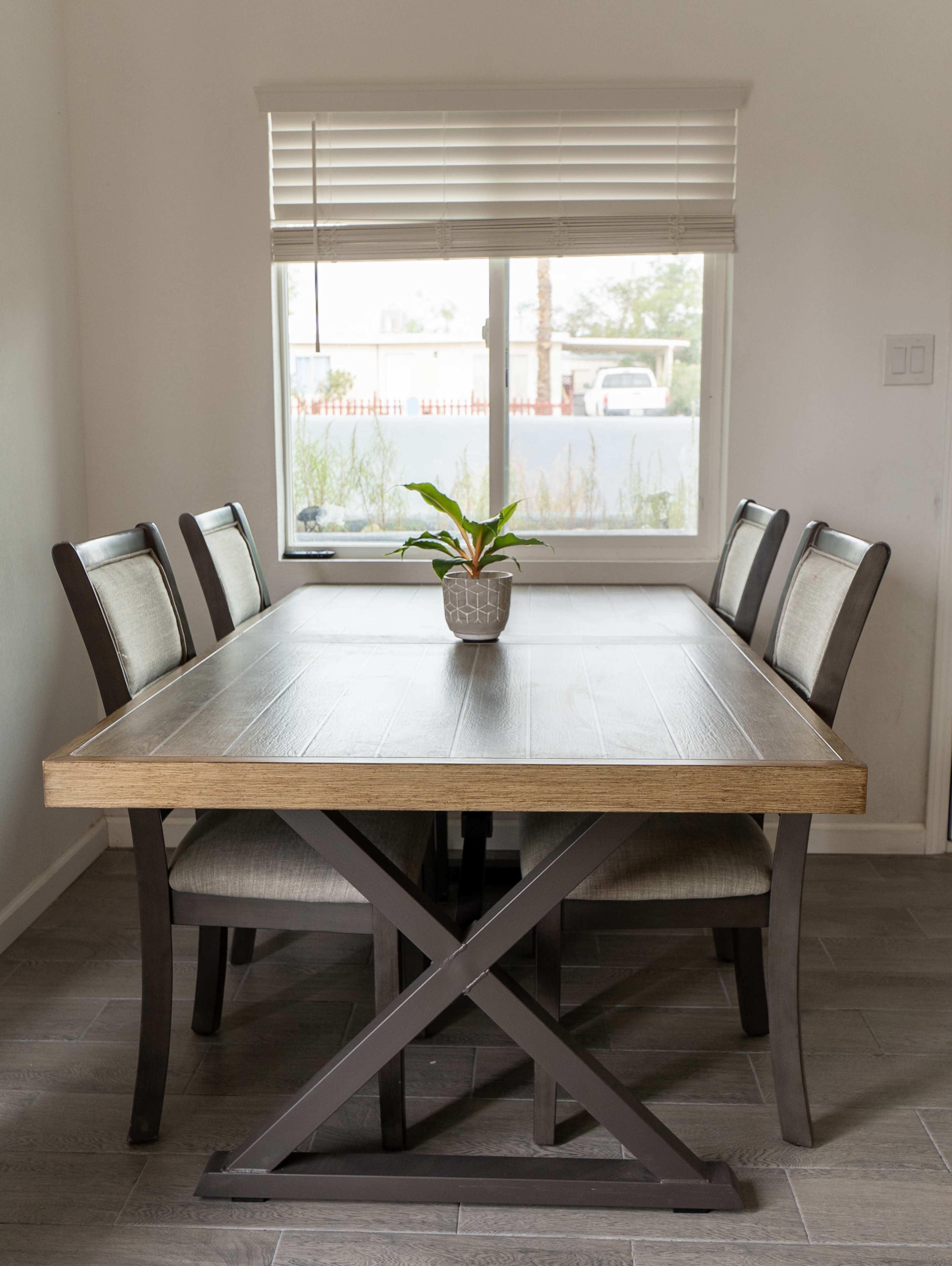 A wooden dining table with four chairs is positioned in front of a window, featuring a small plant in the center.