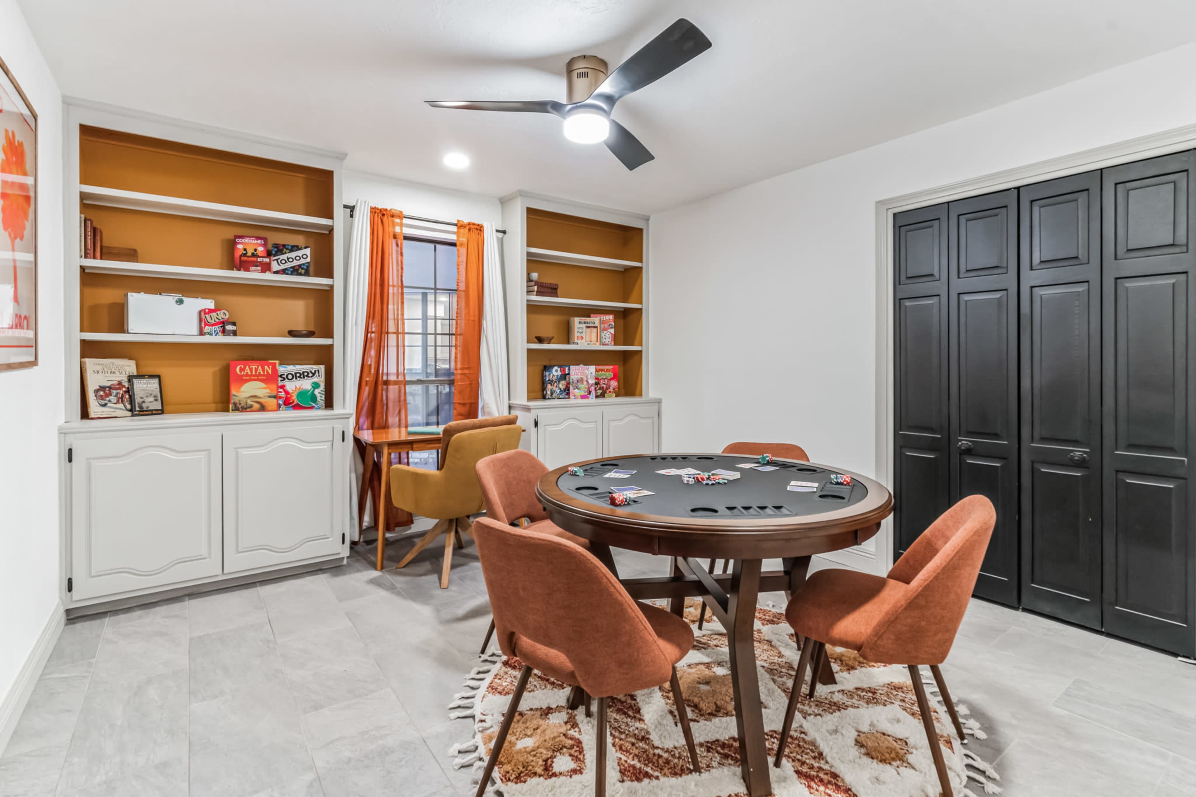 A game room featuring a round table surrounded by four orange chairs, with shelves displaying board games and a window with orange curtains.