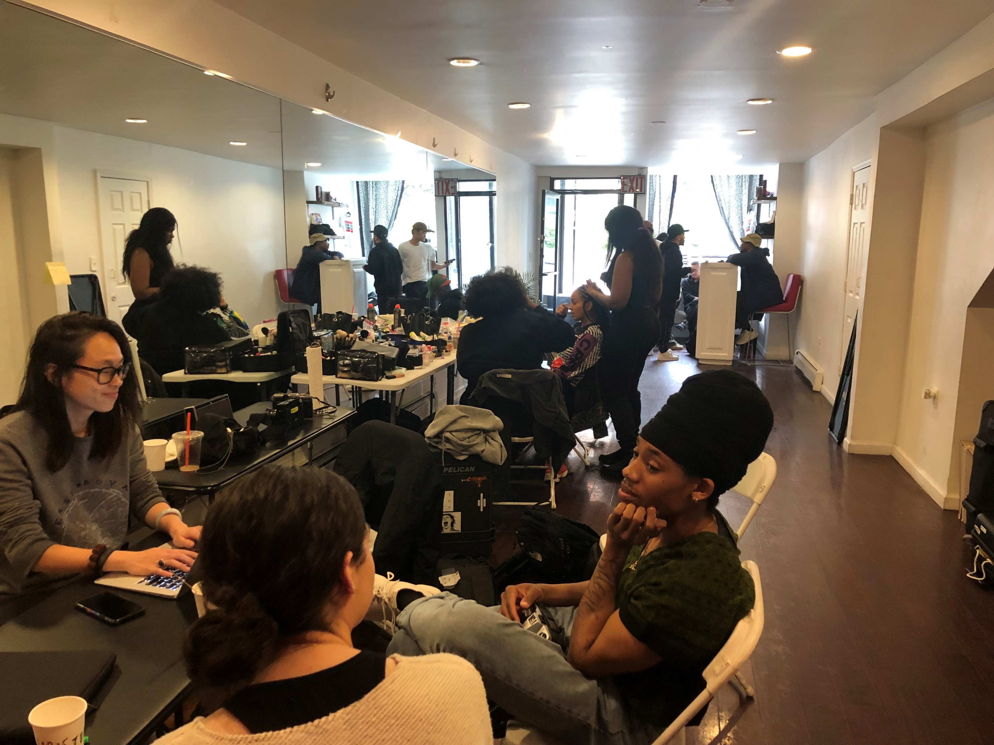A group of people are engaged in hairstyling and conversation in a brightly lit salon with mirrors along one wall.