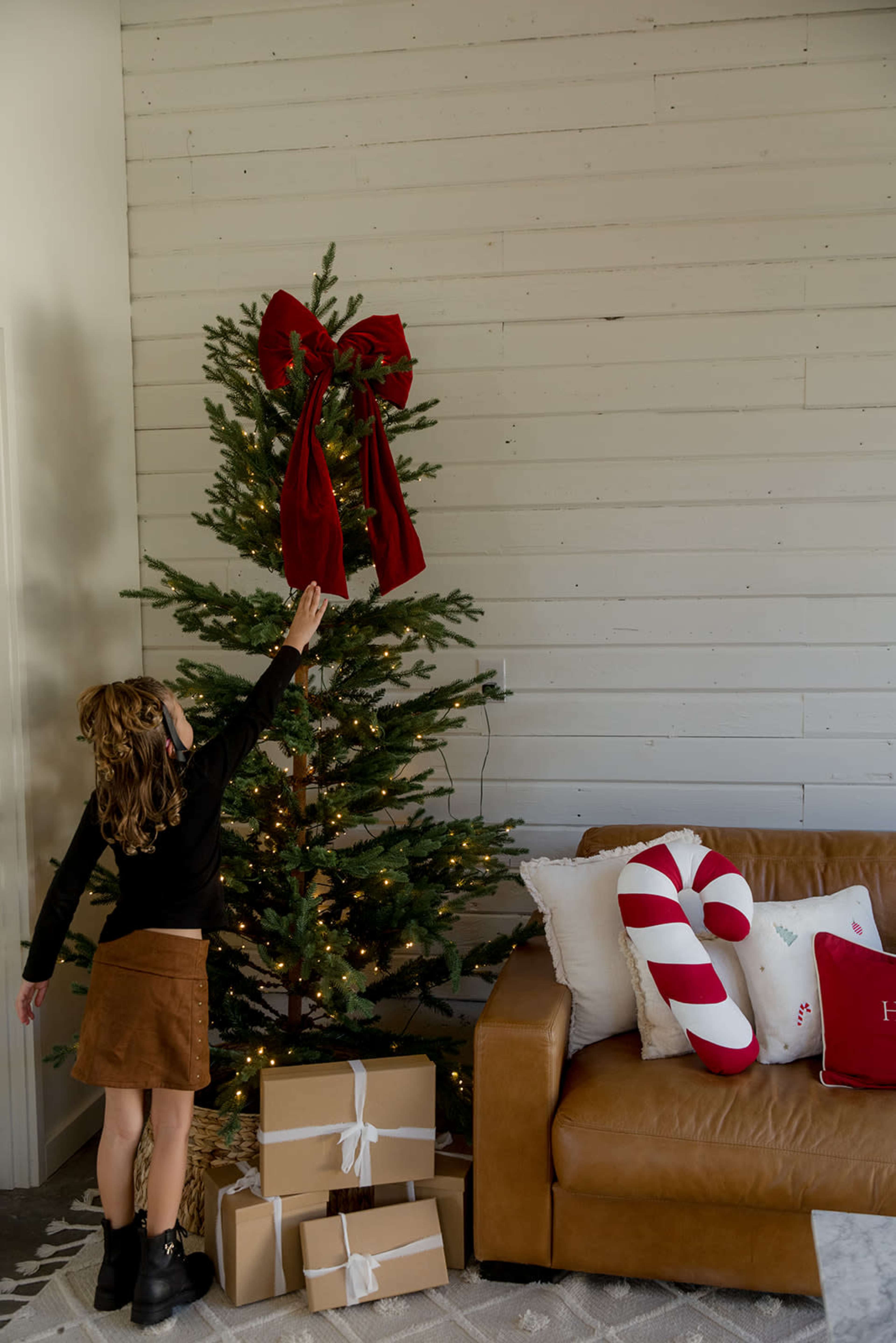 A child reaches towards the top of a Christmas tree decorated with lights and a red bow, while brown gift boxes are placed near a brown couch in a cozy living room.