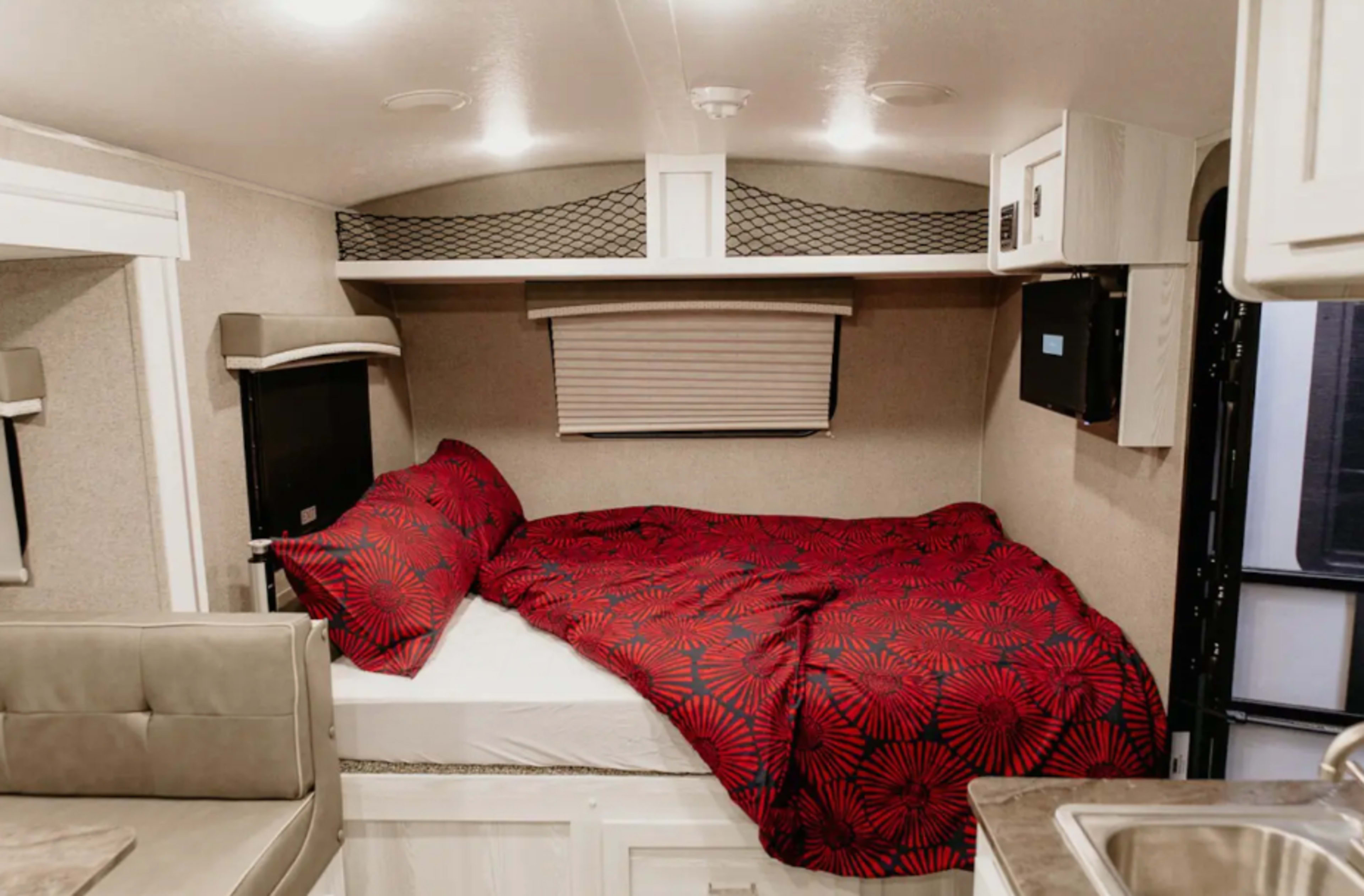 A compact camper interior featuring a bed with a red floral-patterned comforter and a small kitchenette area.