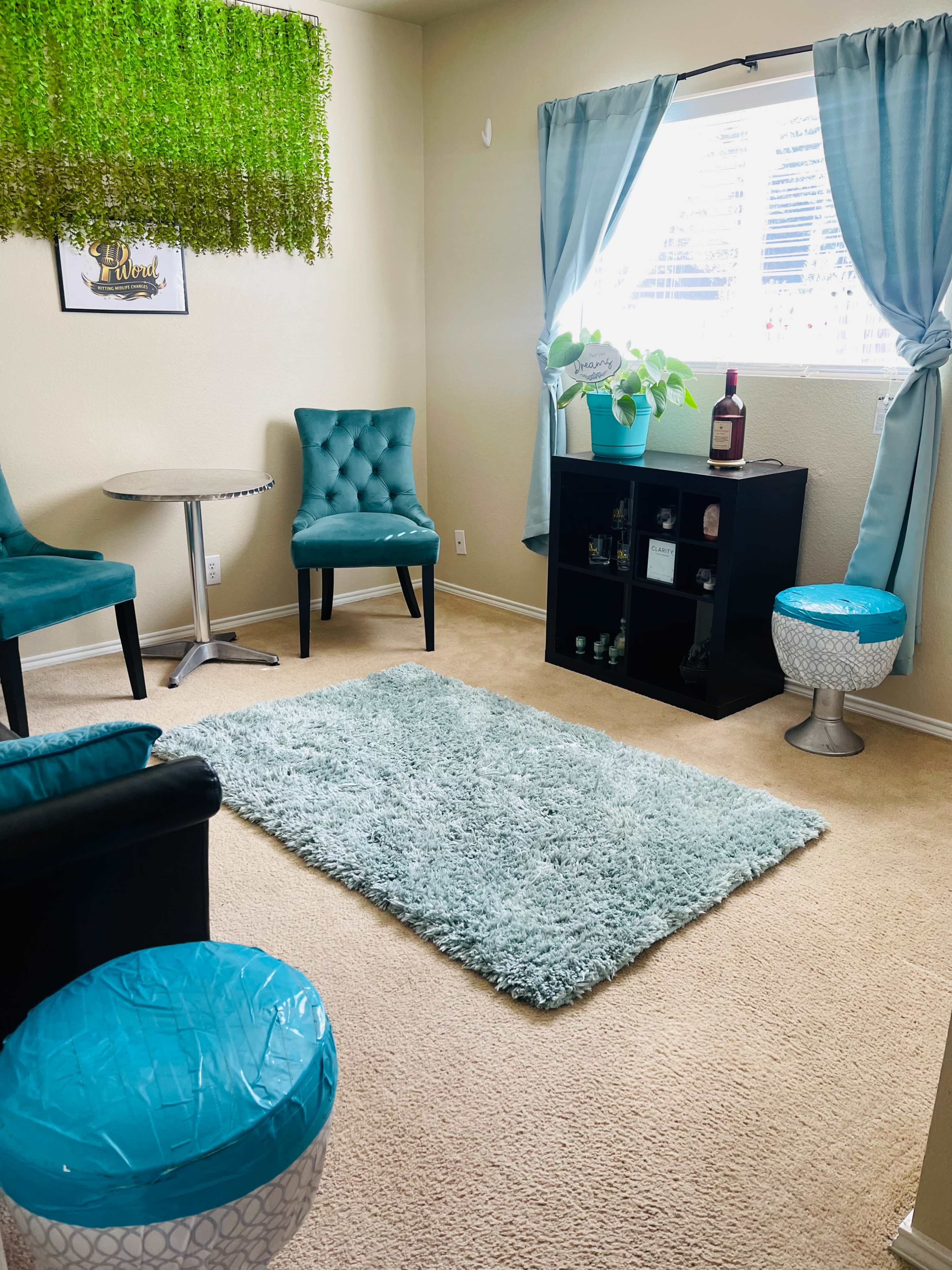 A small seating area features two turquoise chairs, a round table, and a black cabinet, all arranged on a beige carpet with a light blue rug.