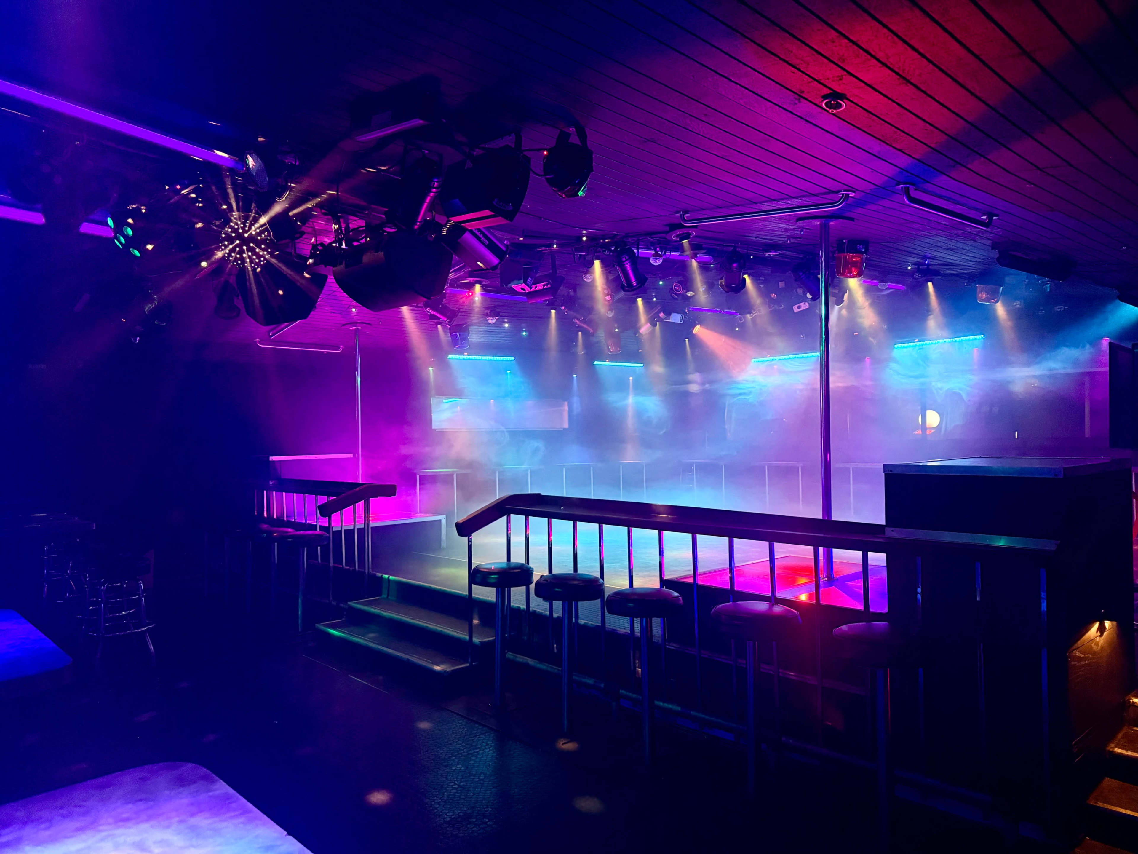 The image shows a dimly lit nightclub with colorful lights, a fog machine, and a raised stage area surrounded by seating.