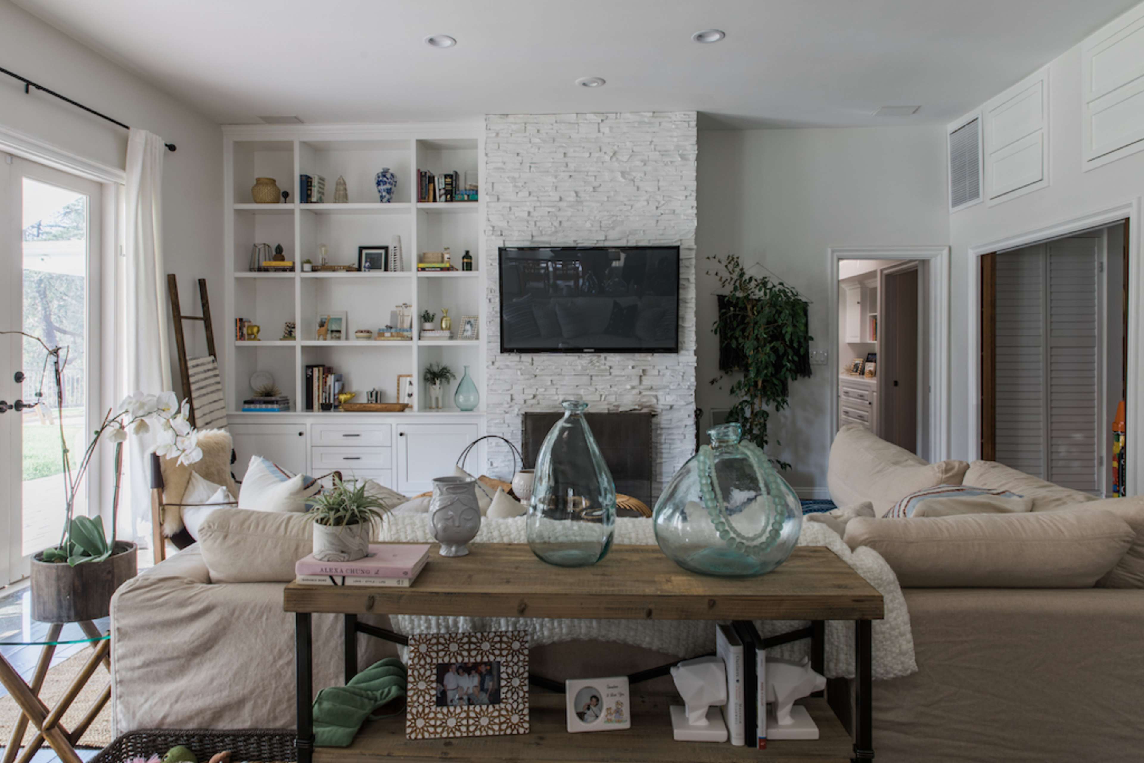 A modern living room features a stone fireplace, shelves filled with decorative items, and a large sectional sofa facing a mounted television.