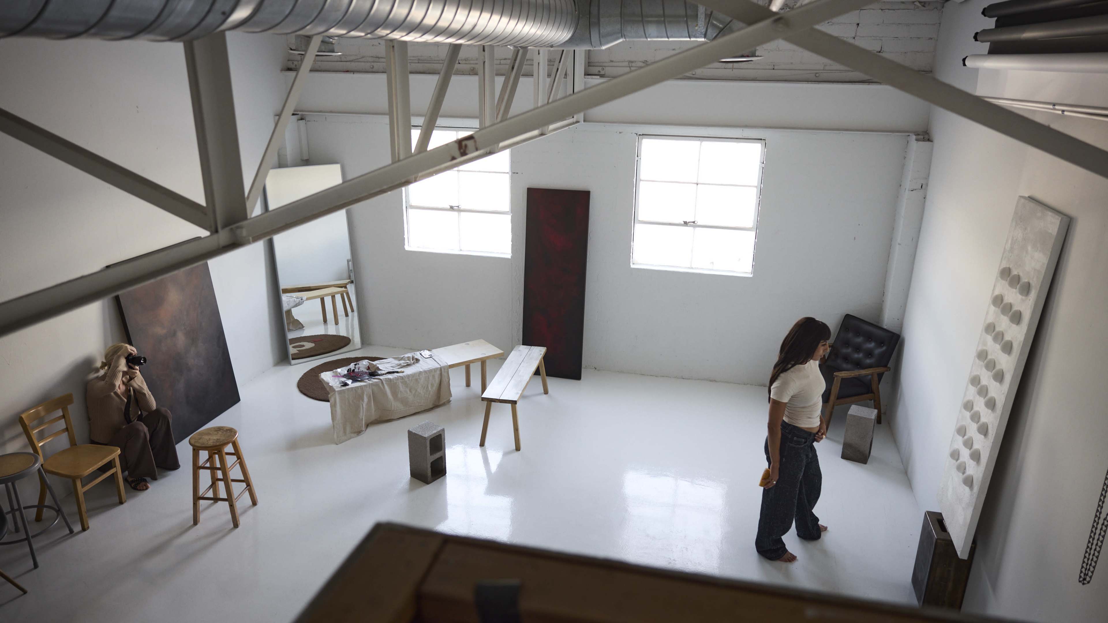 The image shows a spacious, minimalist art studio with a woman walking across the floor, surrounded by various art pieces and furniture.