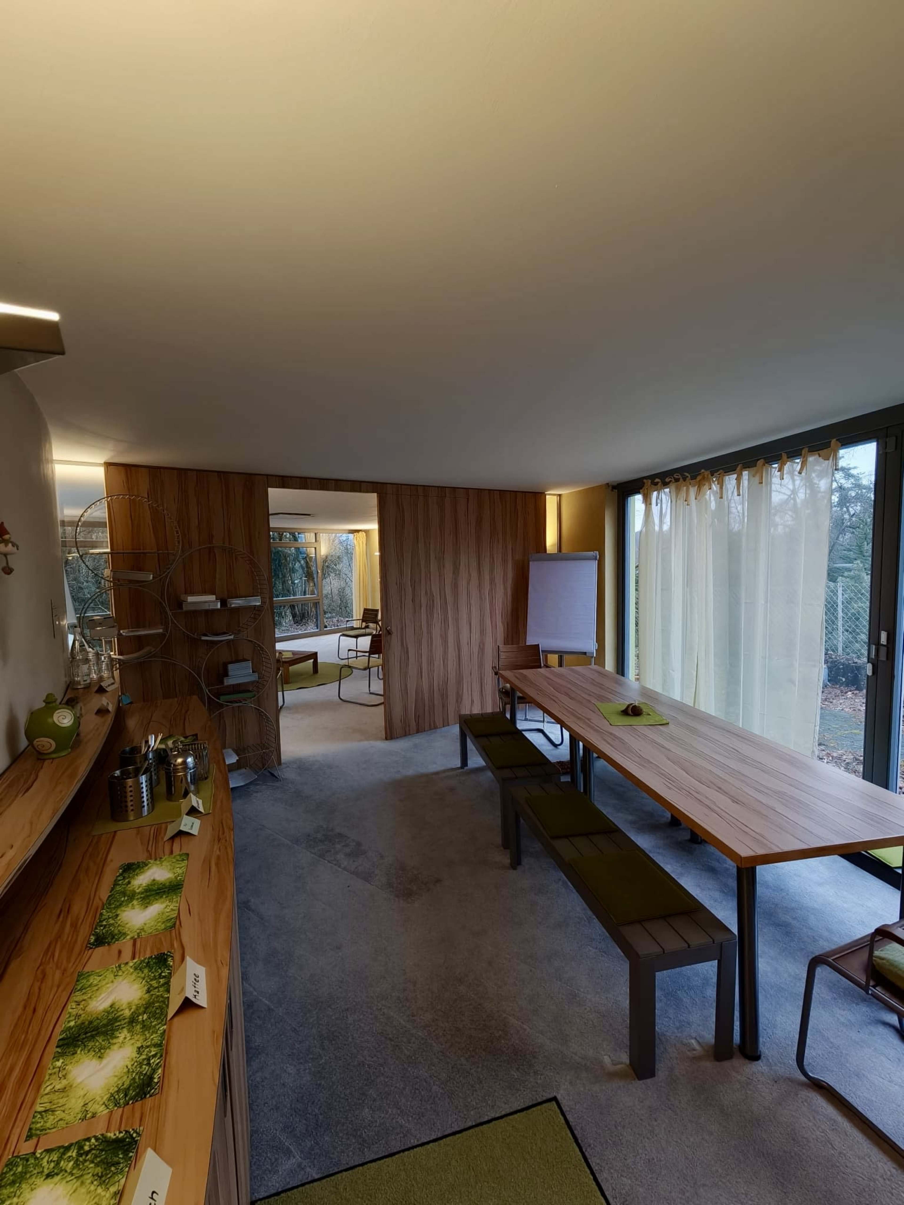 The image shows a modern interior space featuring a long wooden table, benches, and a shelf with various items, along with large windows allowing natural light to enter.