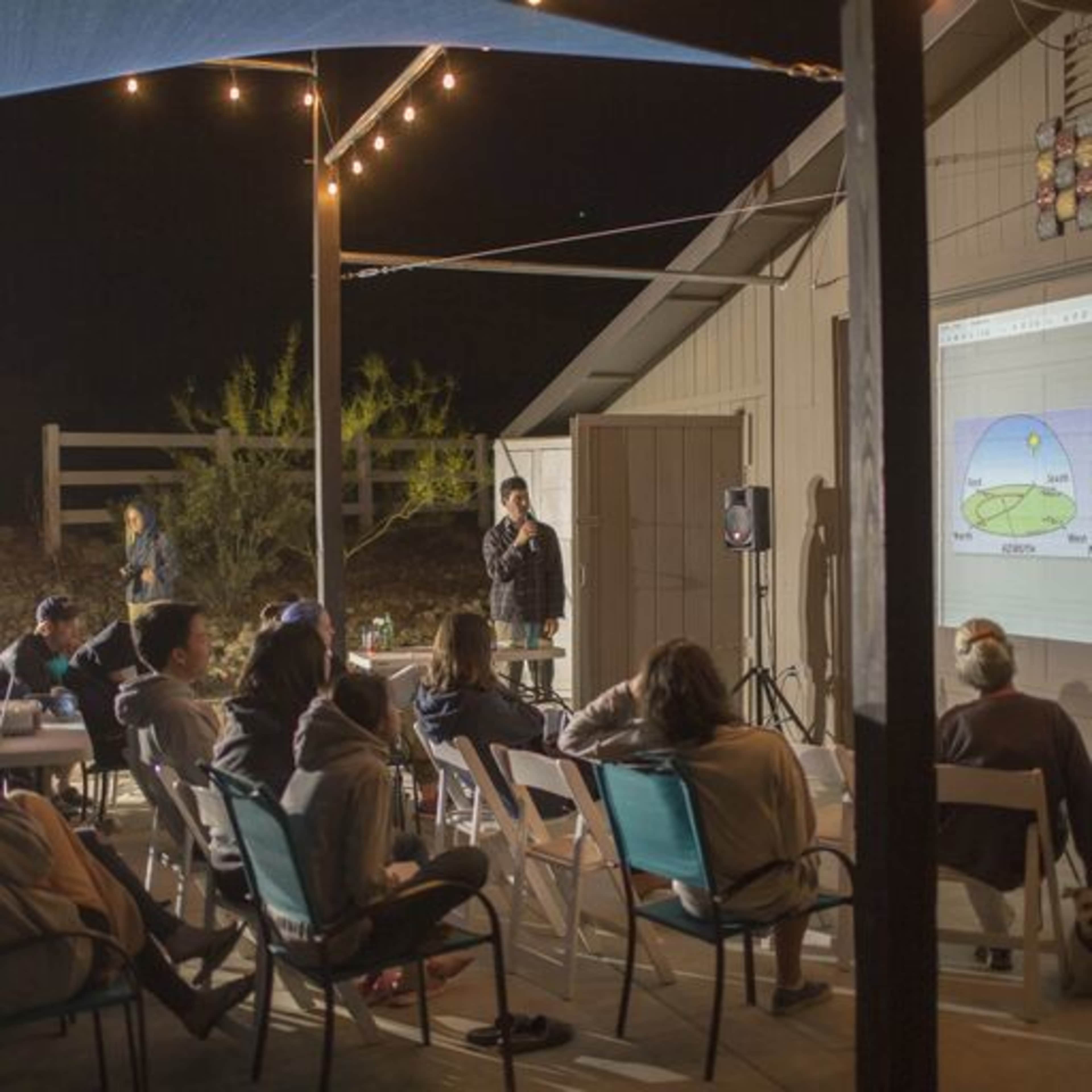 A group of people is seated outdoors, watching a presentation projected onto a wall beside a building at night.
