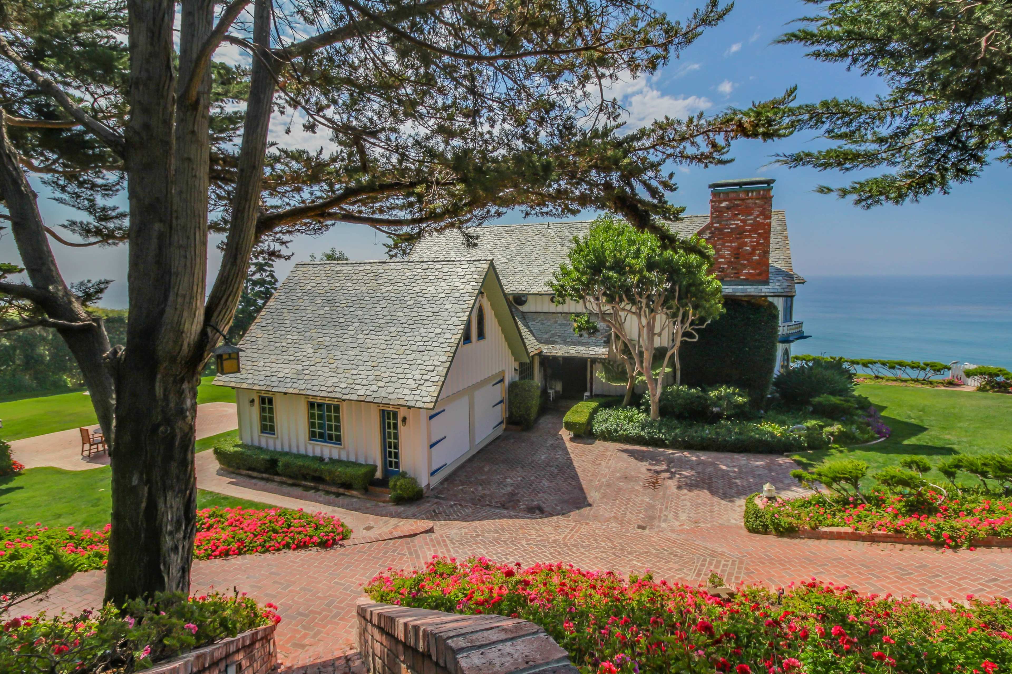 A large coastal house with a shingled roof is surrounded by lush greenery and flowers, overlooking the ocean.