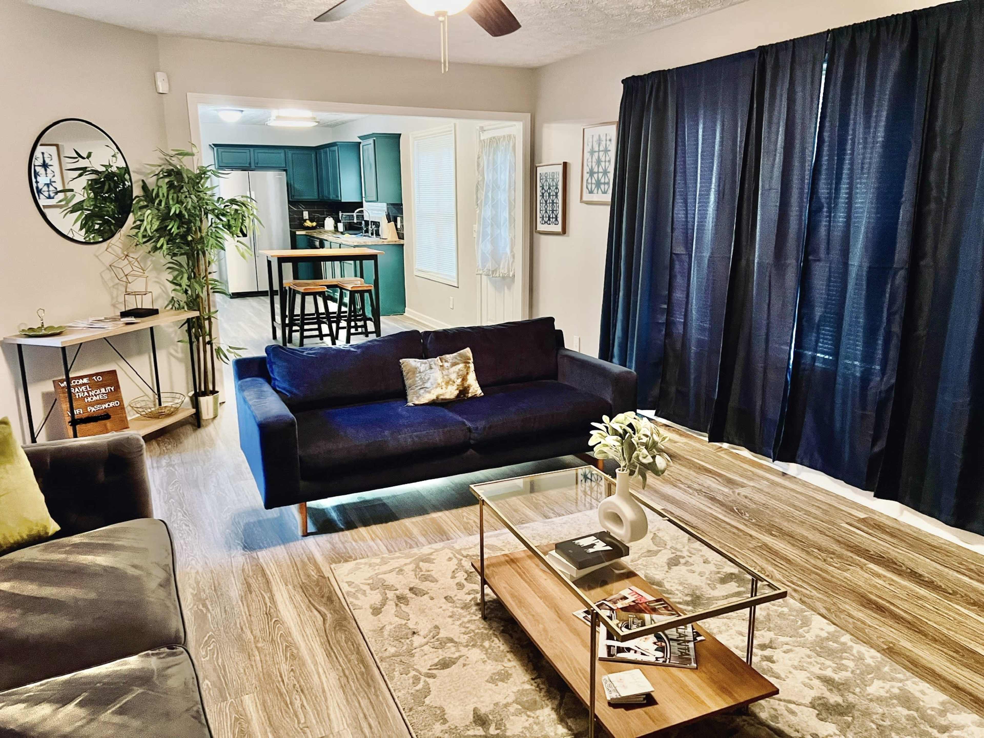 The image shows a cozy living room with a dark blue sofa, a glass coffee table on a patterned rug, and a doorway leading to a kitchen with teal cabinets.