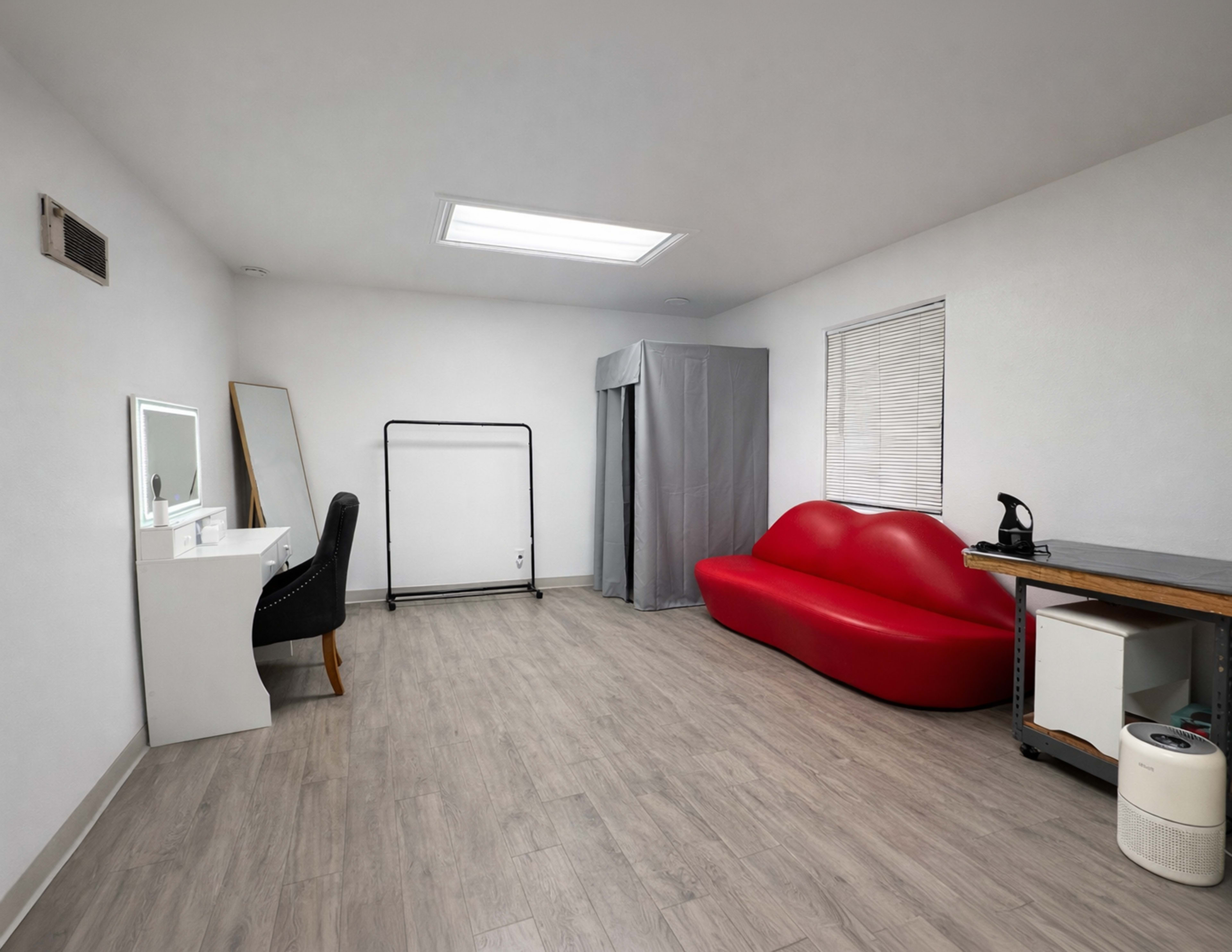 The image shows a minimalist room featuring a red seating area, a dressing mirror, a clothing rack, and a gray privacy curtain.