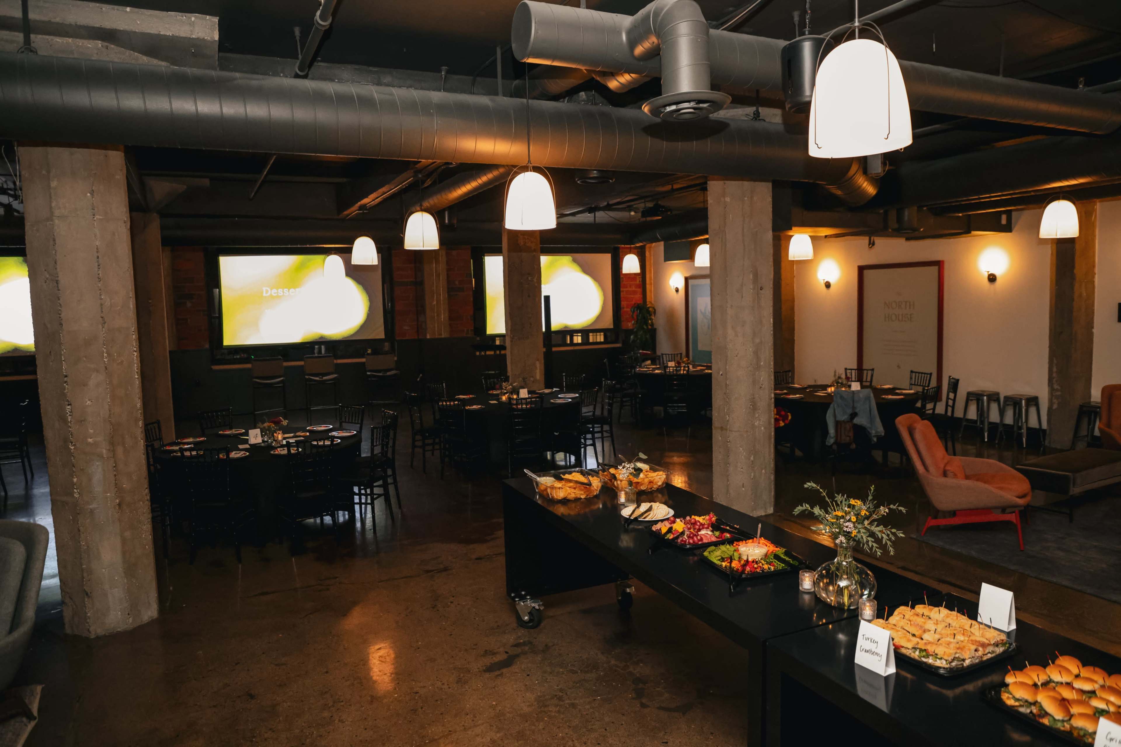 The image shows an event space with dining tables, a buffet table displaying food, and two screens on the walls.