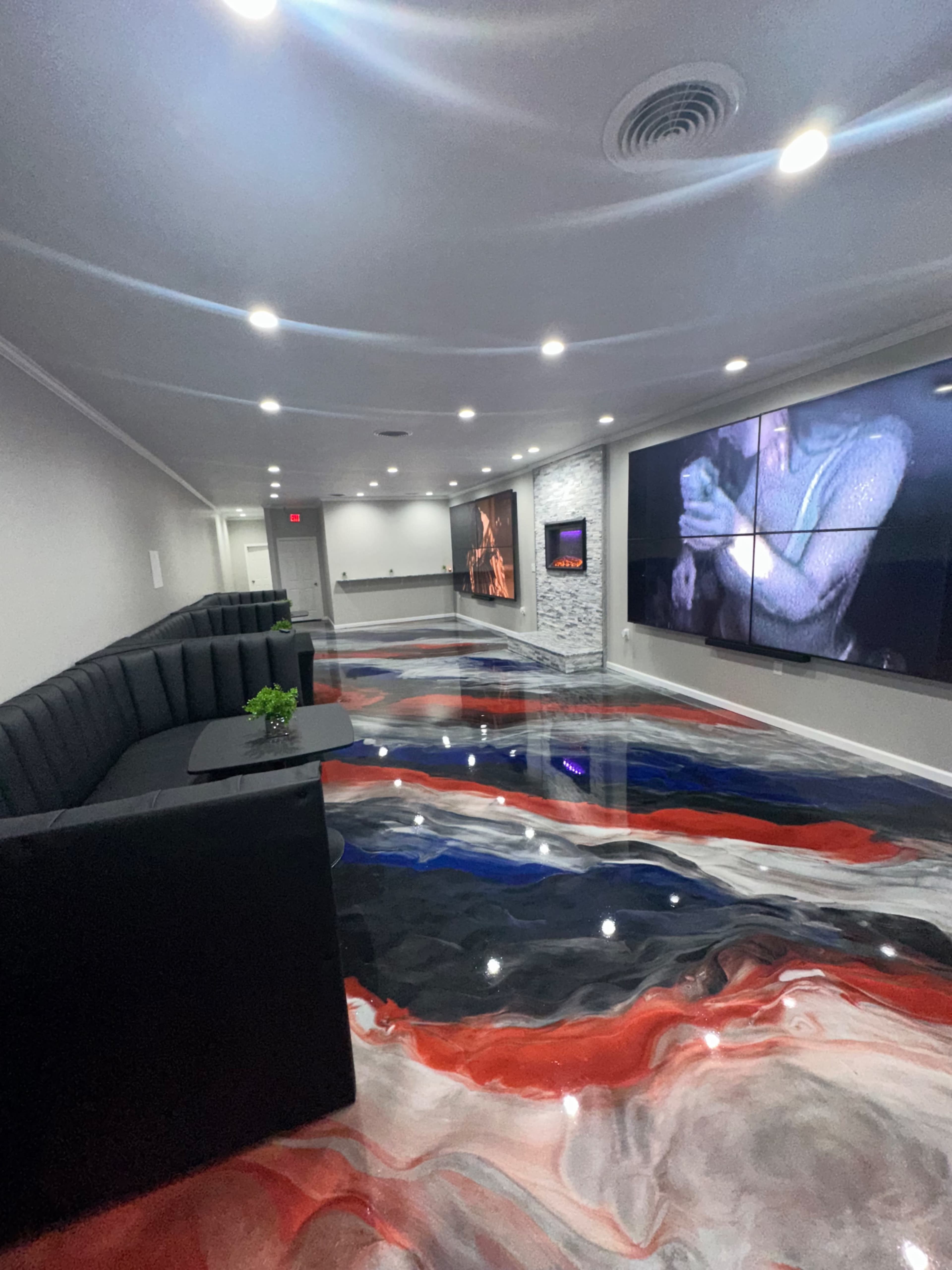 The image shows a modern hallway with a glossy, swirling red, white, and blue floor, black seating along the walls, and large screens displaying images.