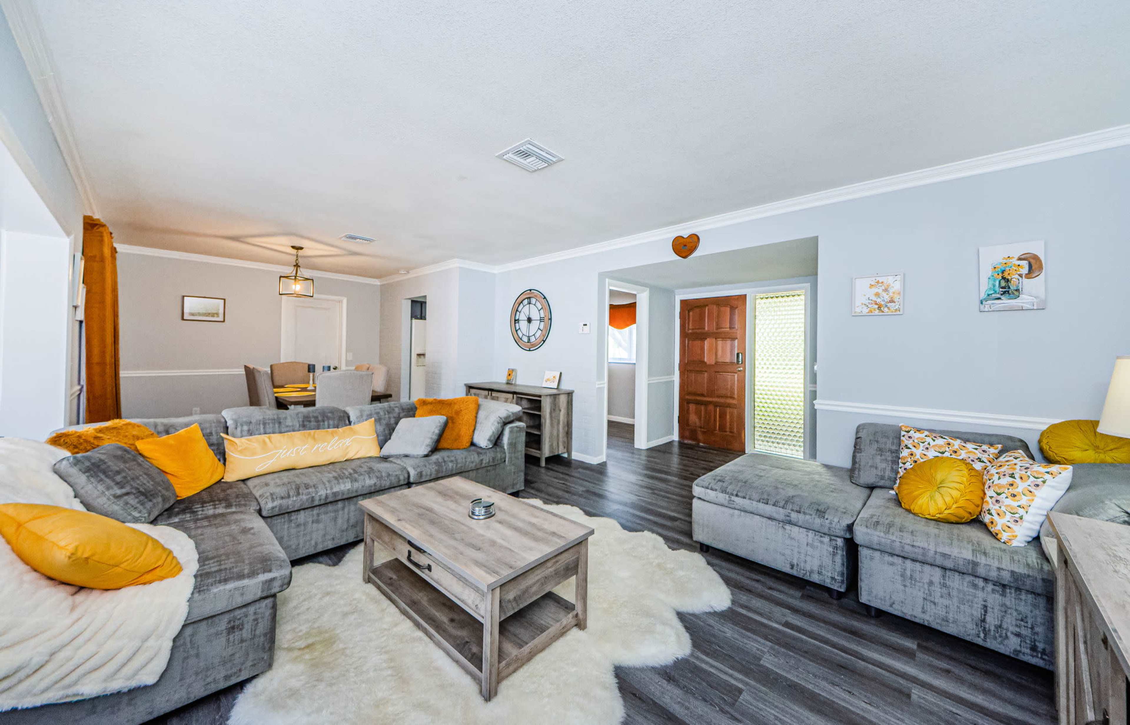 The living room features gray furniture with orange accent pillows, a wooden coffee table, and a light-colored area rug, illuminated by natural light from a nearby door.