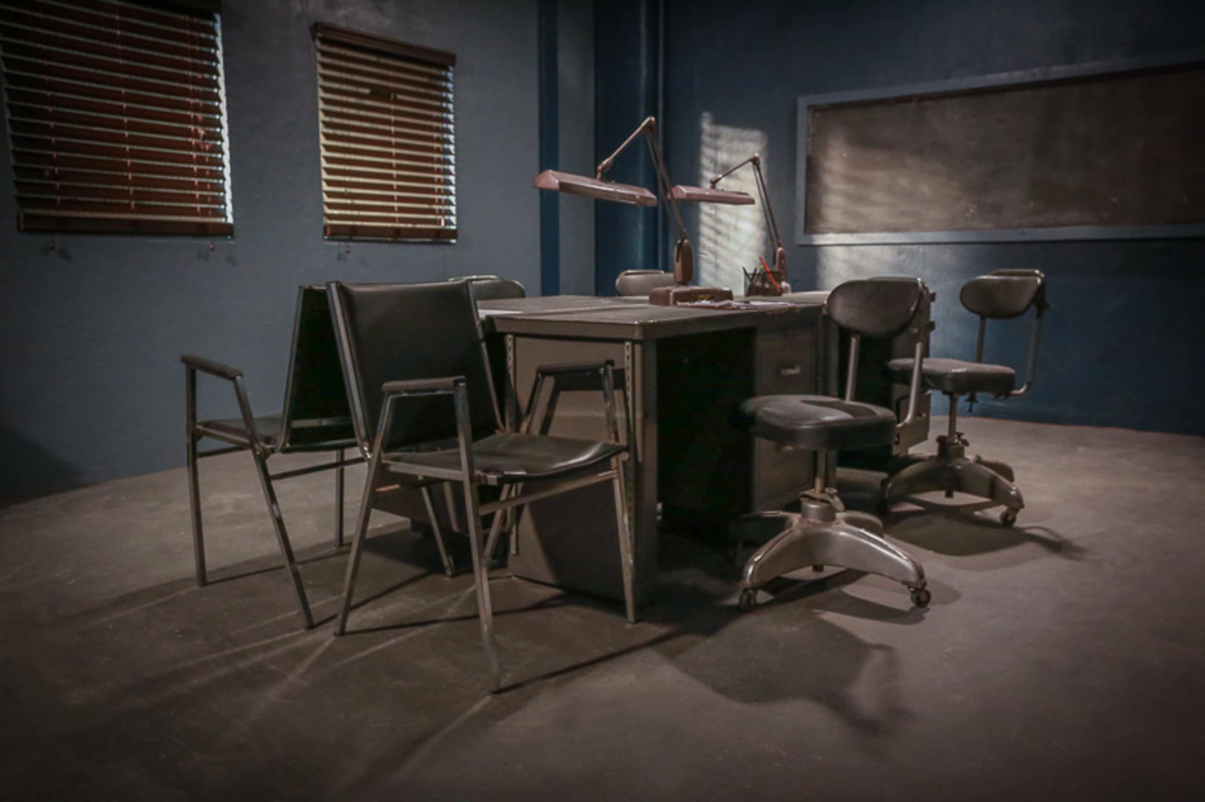 A dimly lit room features a table surrounded by several metal chairs and a couple of task chairs, with wooden blinds casting shadows on the walls.