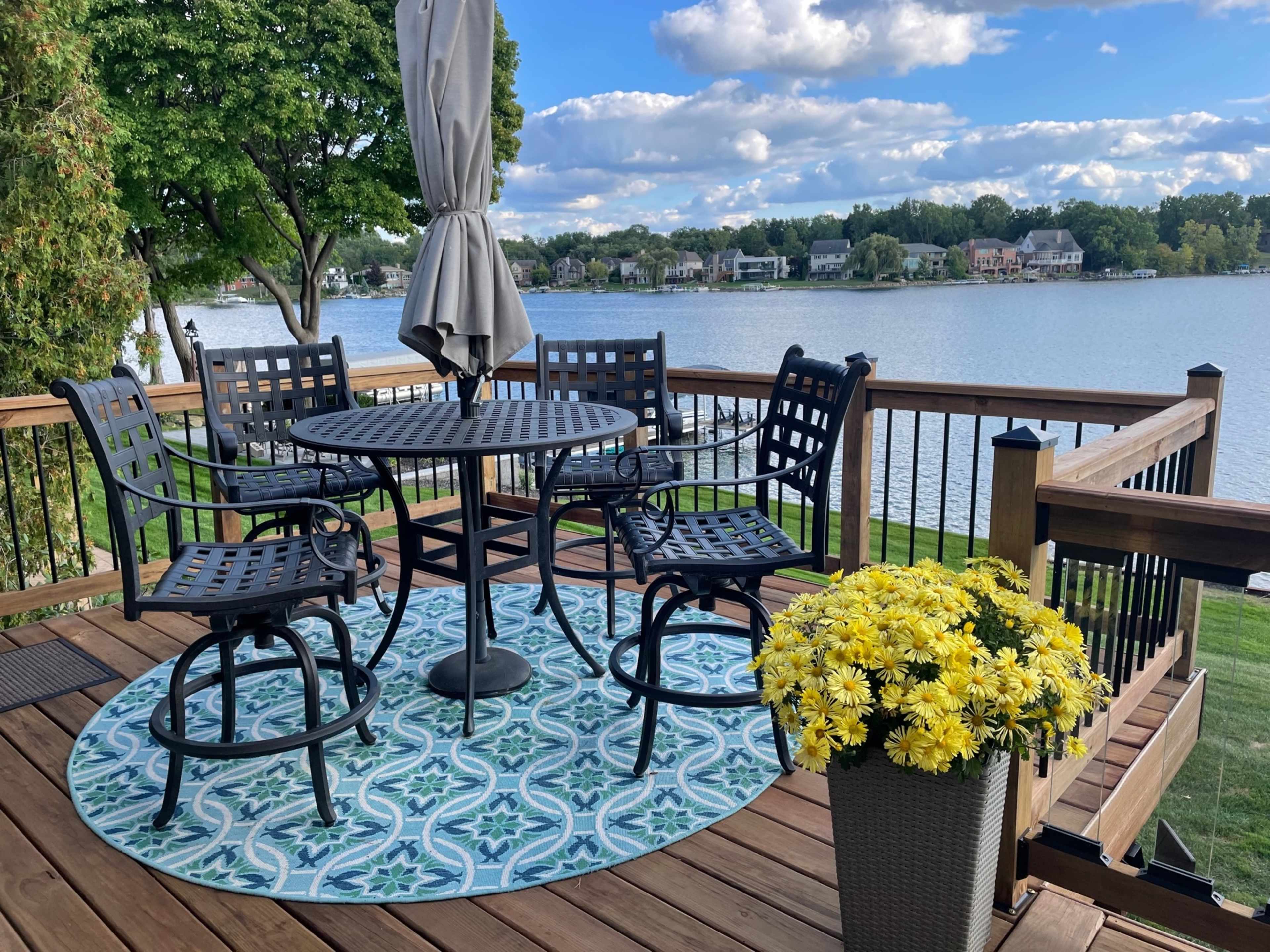 A round table with four chairs is set on a wooden deck overlooking a lake, surrounded by greenery and a potted plant filled with yellow flowers.