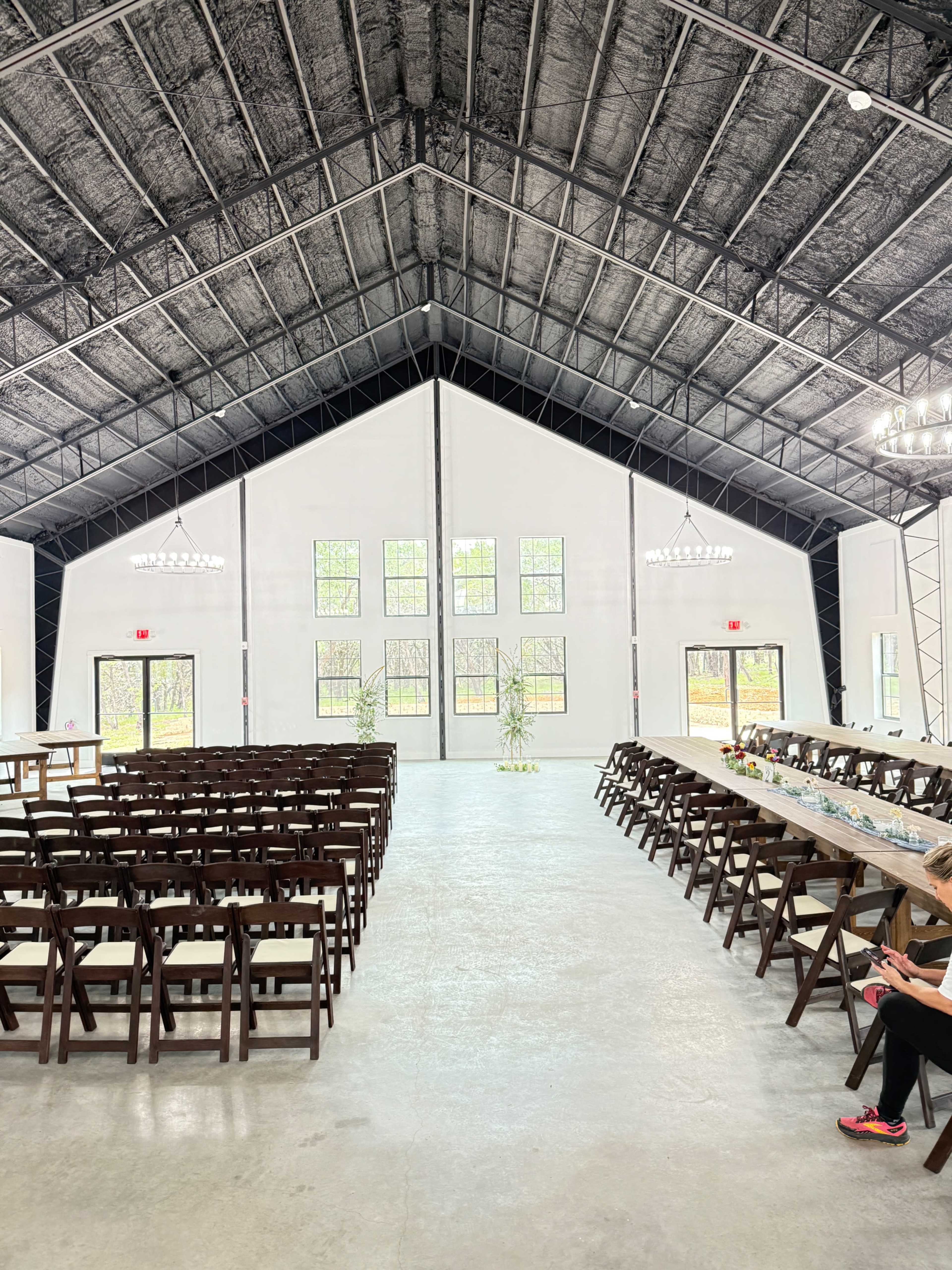 The image shows a spacious indoor event venue with rows of wooden chairs facing a long table, set against a backdrop of large windows.