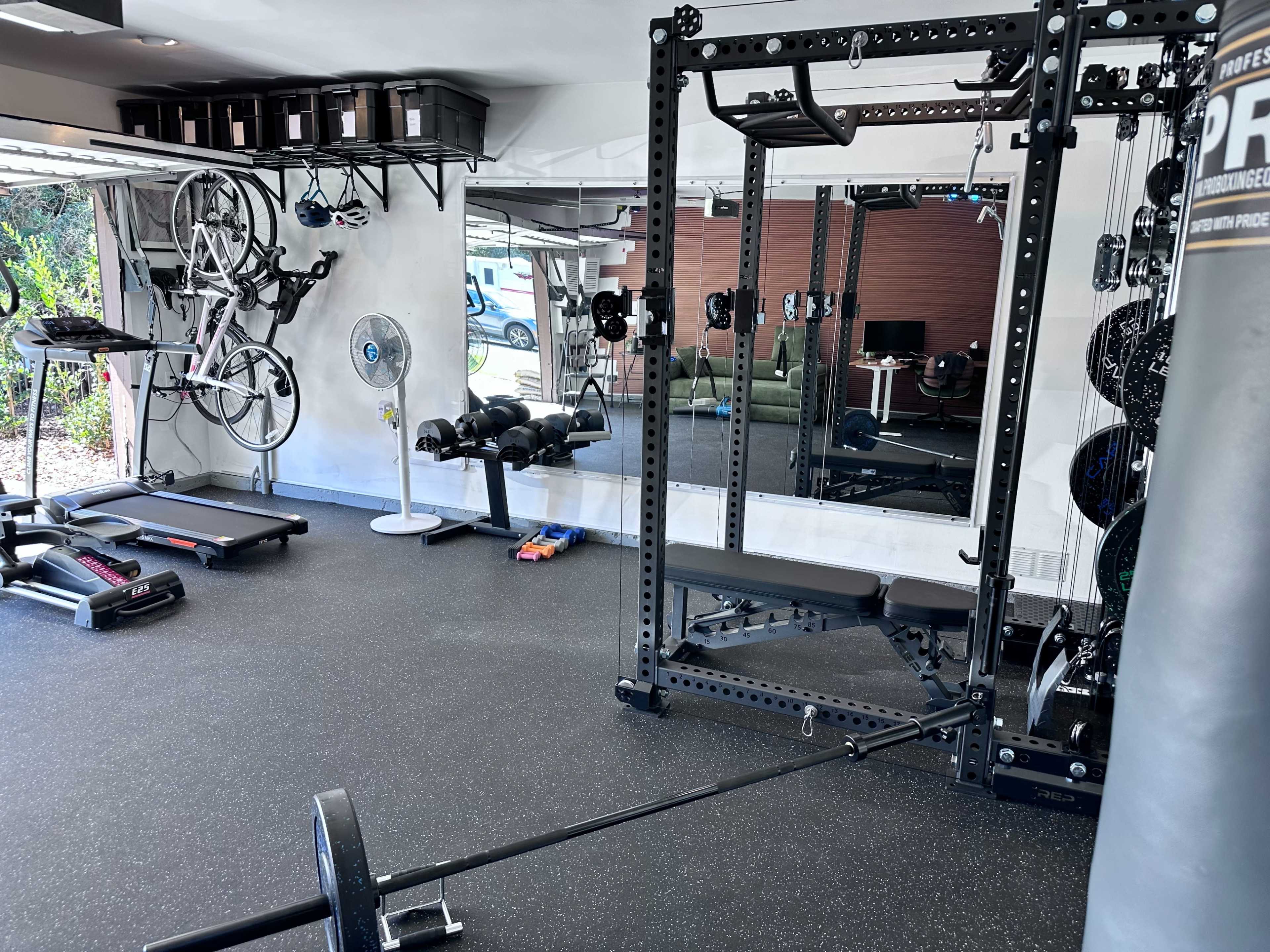 The image shows a well-equipped home gym featuring a squat rack, dumbbells, a treadmill, and exercise bicycles mounted on the wall.